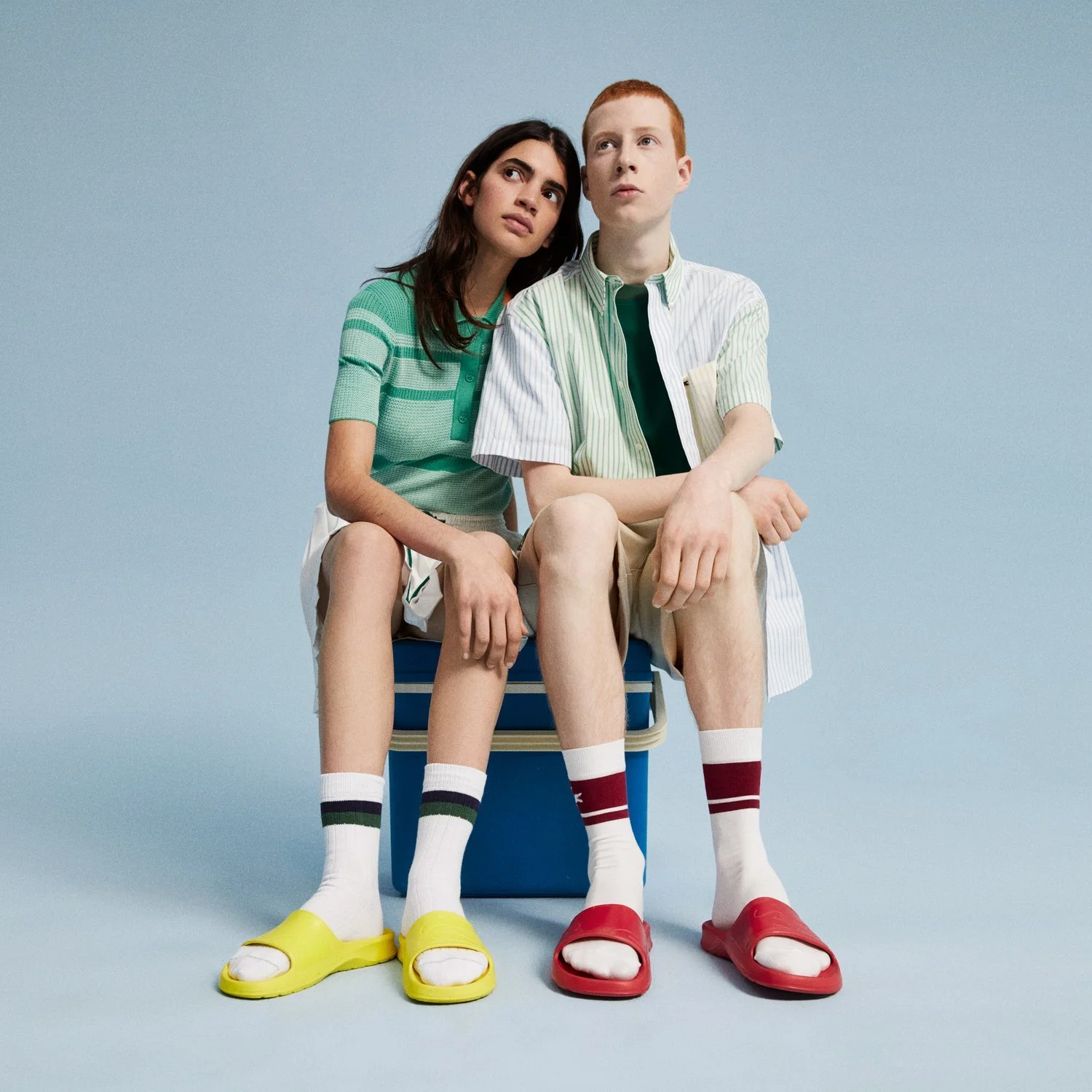 A boy and girl sit close together on a blue bench against a light blue background, wearing casual summer clothes and colorful sandals.