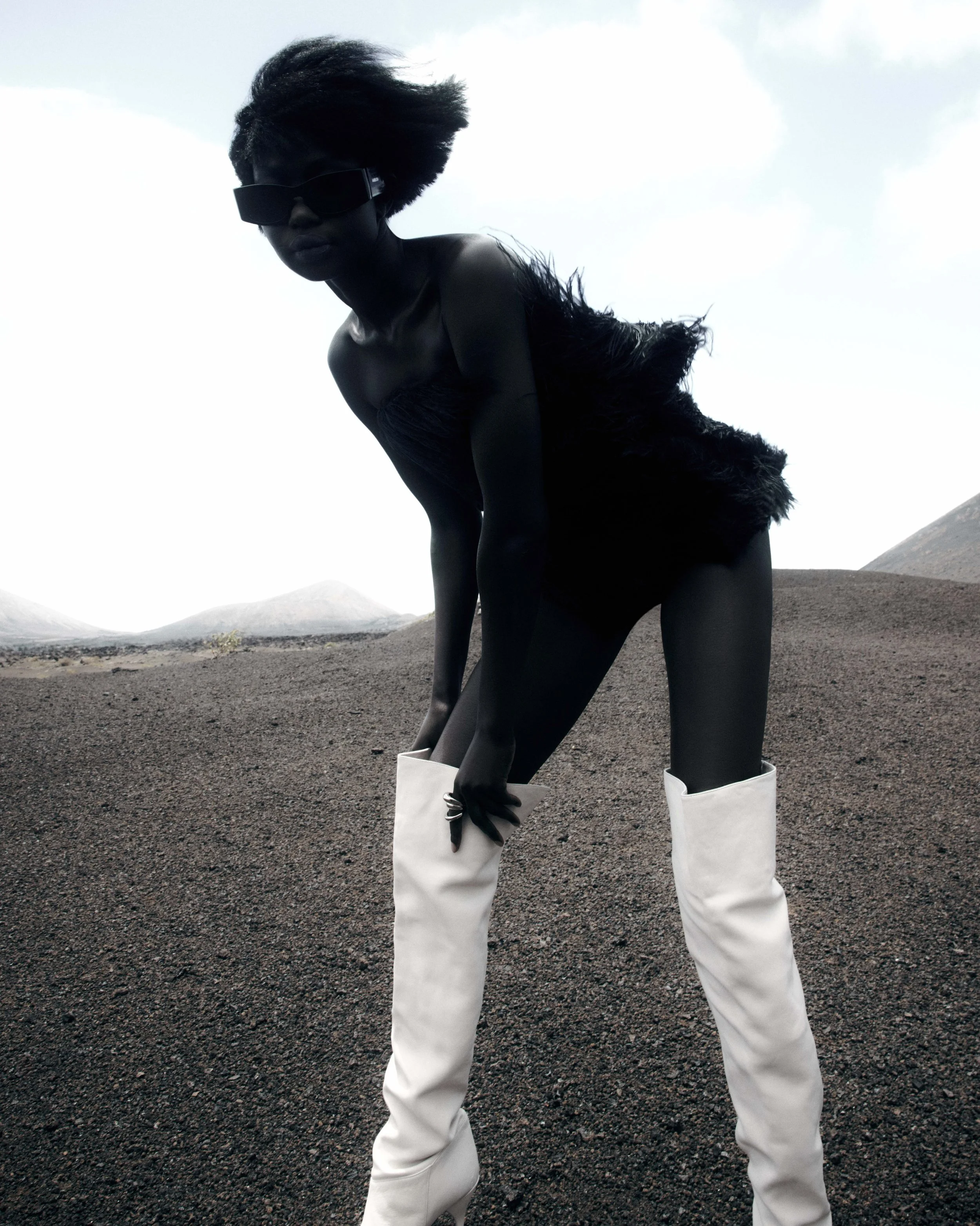 A person with dark skin, black hair, wearing large black sunglasses, a black strapless dress with feather details, and white knee-high boots, is posed outdoors on a landscape with dark soil or volcanic ash and mountainous terrain in the background.