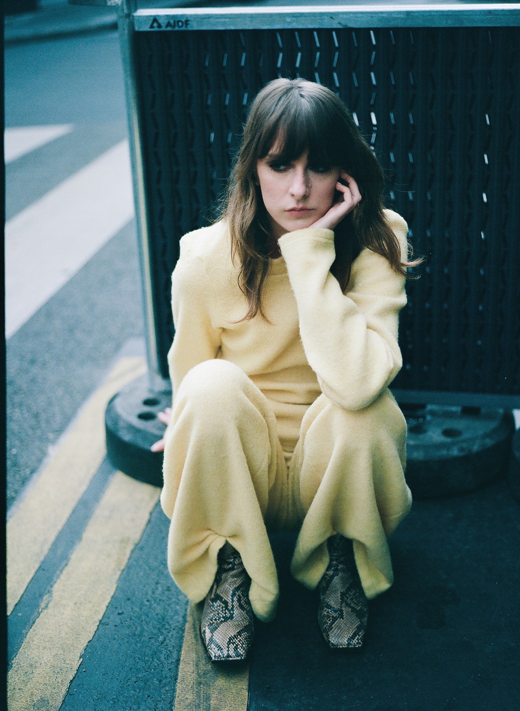 A young woman in a yellow outfit sitting on a road near a black tire, resting her head on her hand and looking away.