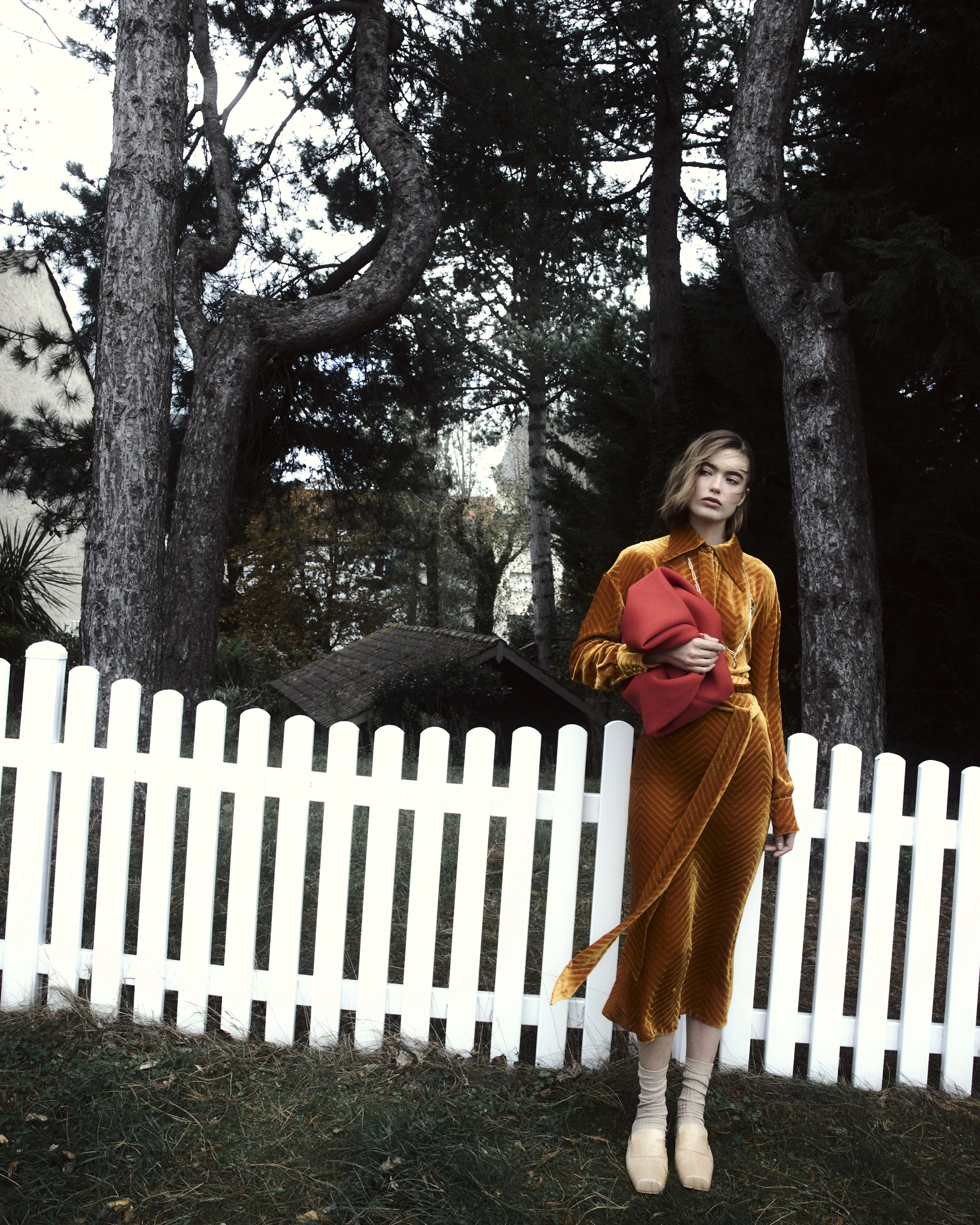 A woman standing in front of a white picket fence, wearing a rusty orange outfit, holding a red bag, with trees and a small wooden shed in the background.