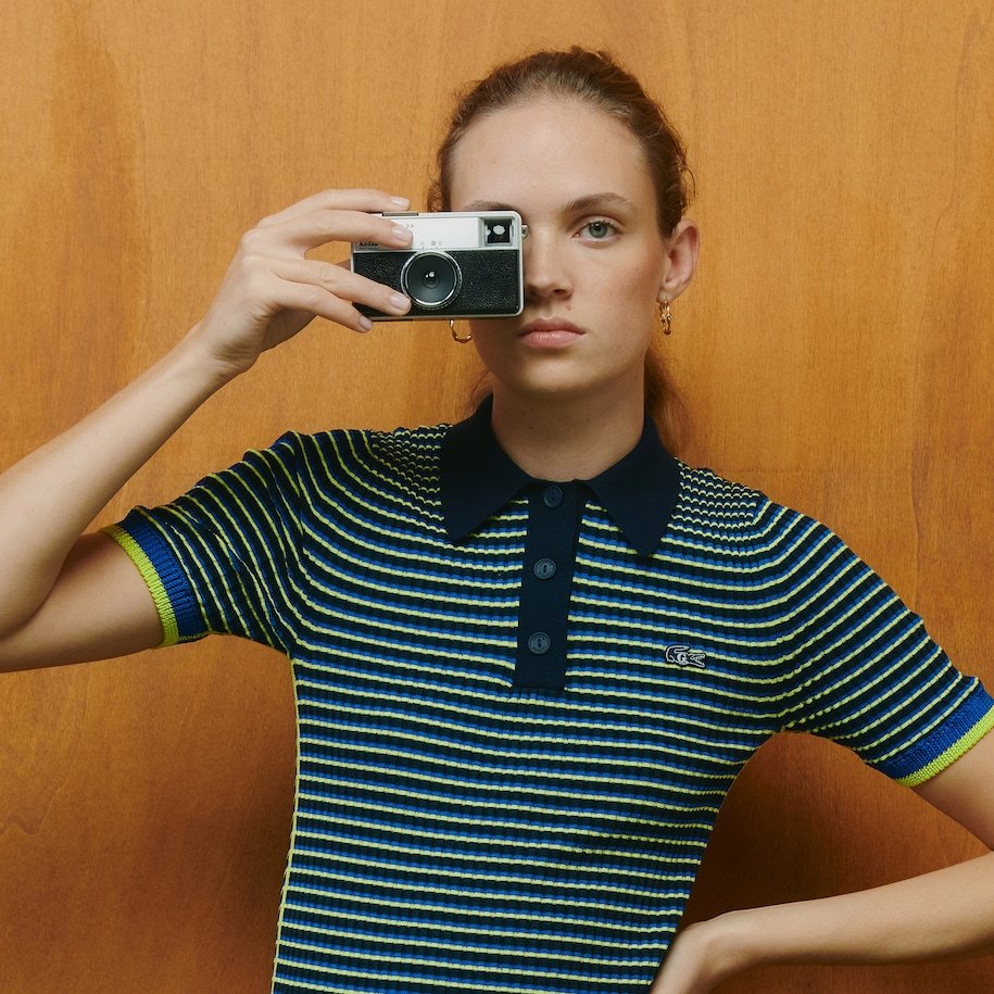 A young woman taking a selfie in a mirror with a vintage camera, wearing a multicolored striped polo shirt, standing against a wooden background.