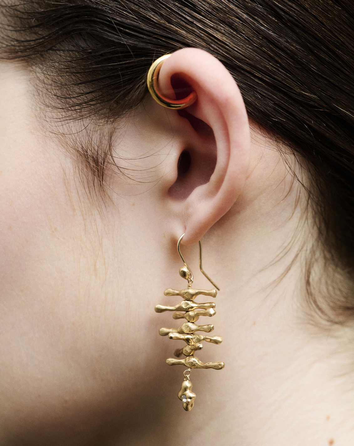 Close-up of a person’s ear with a gold hoop earring and a large, abstract gold dangle earring.