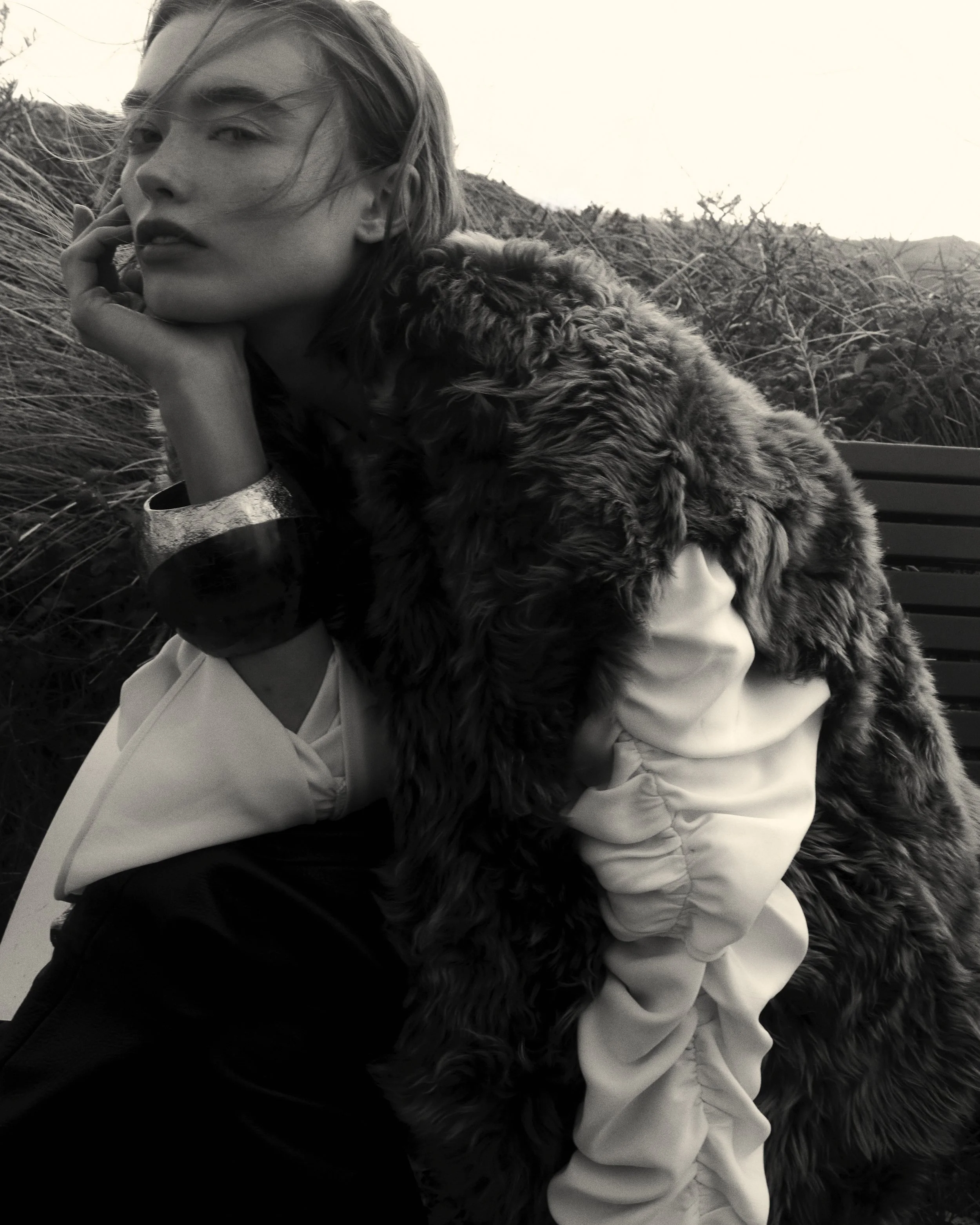 Black and white photo of a woman with short hair, resting her chin on her hand, wearing a layered outfit with a fur coat and a bracelet, sitting outdoors with grass and hills in the background.