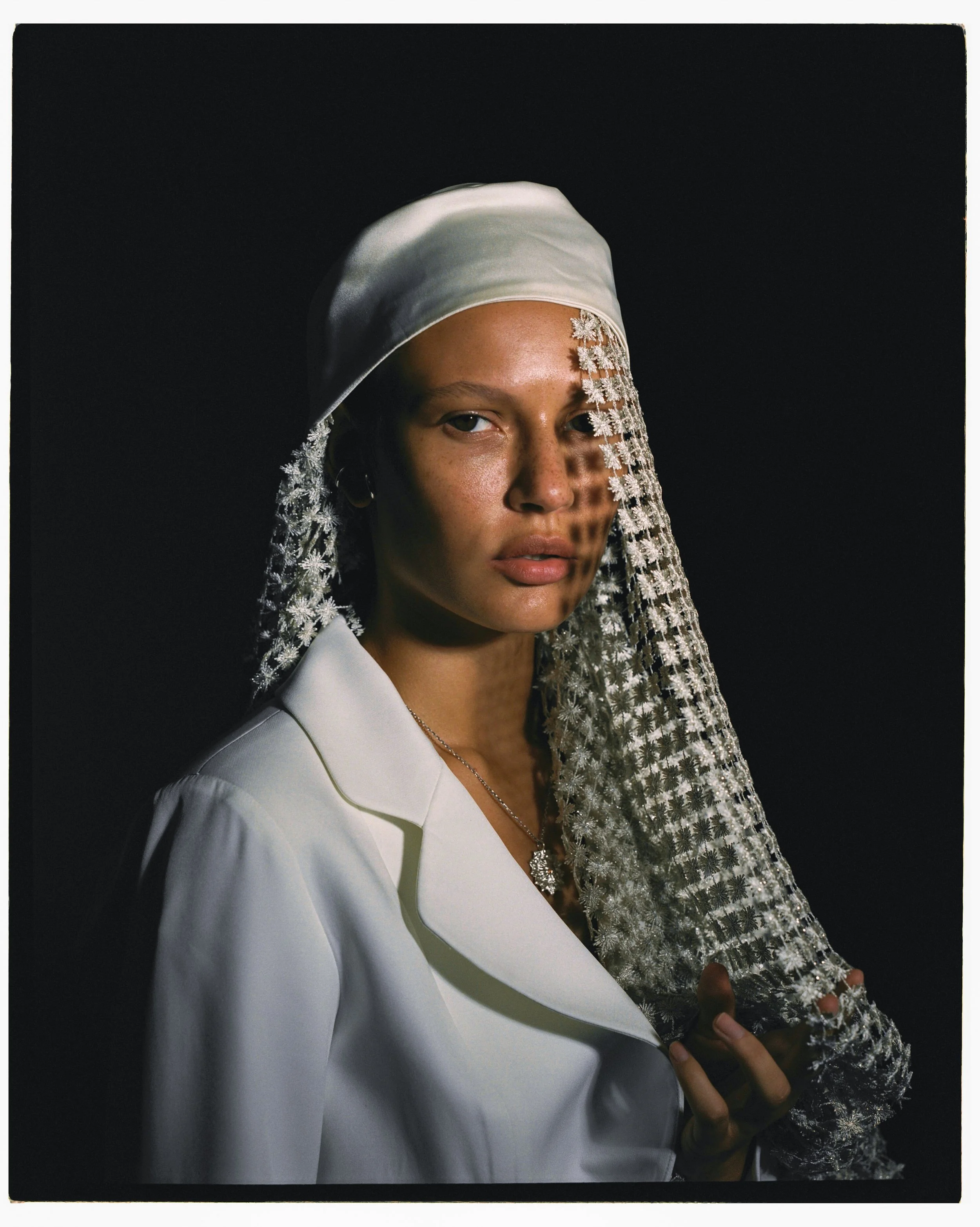 A woman in a white shirt with a white headscarf and lace veil, holding the veil with one hand, standing against a dark background with a shadow pattern on her face.