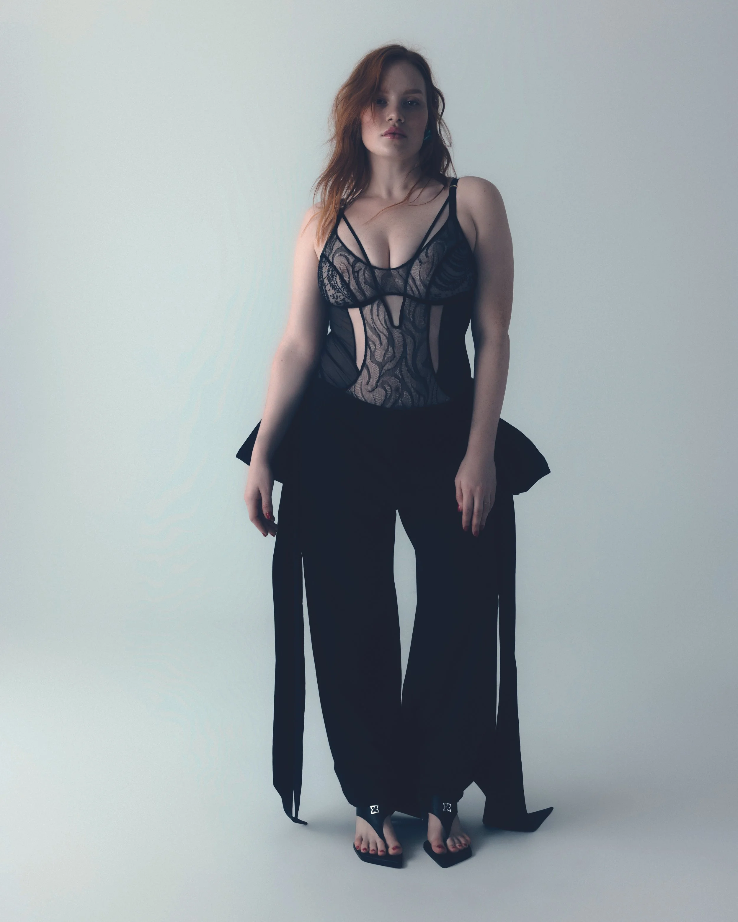 A woman with red hair wearing a black lace bodysuit and loose black pants standing against a light-colored background.