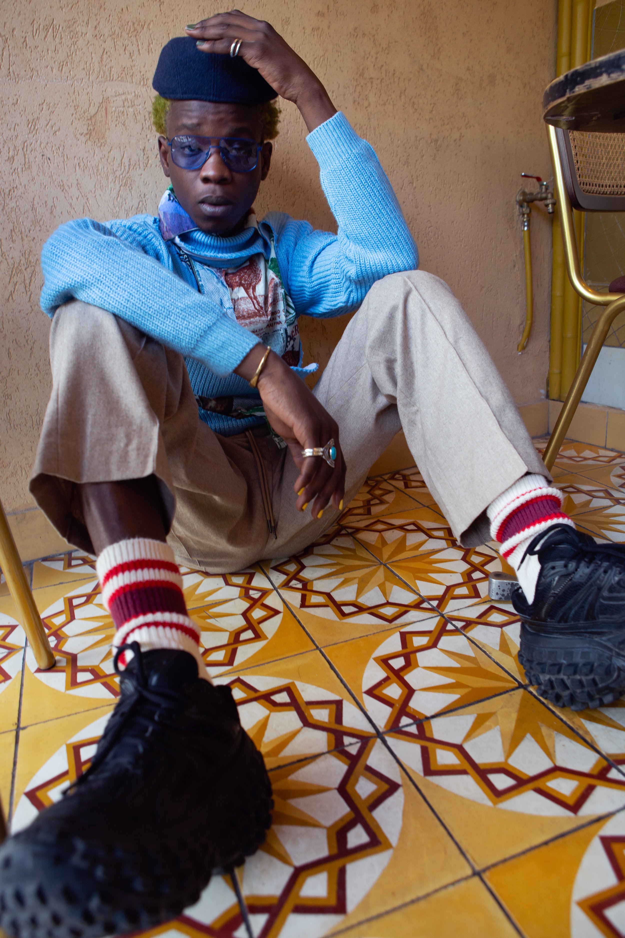 A young person with curly blonde hair, wearing glasses, a black cap, a blue sweater, beige pants, red and white striped socks, and black shoes, sitting on a patterned tile floor, leaning against a beige wall, with individual chairs nearby.