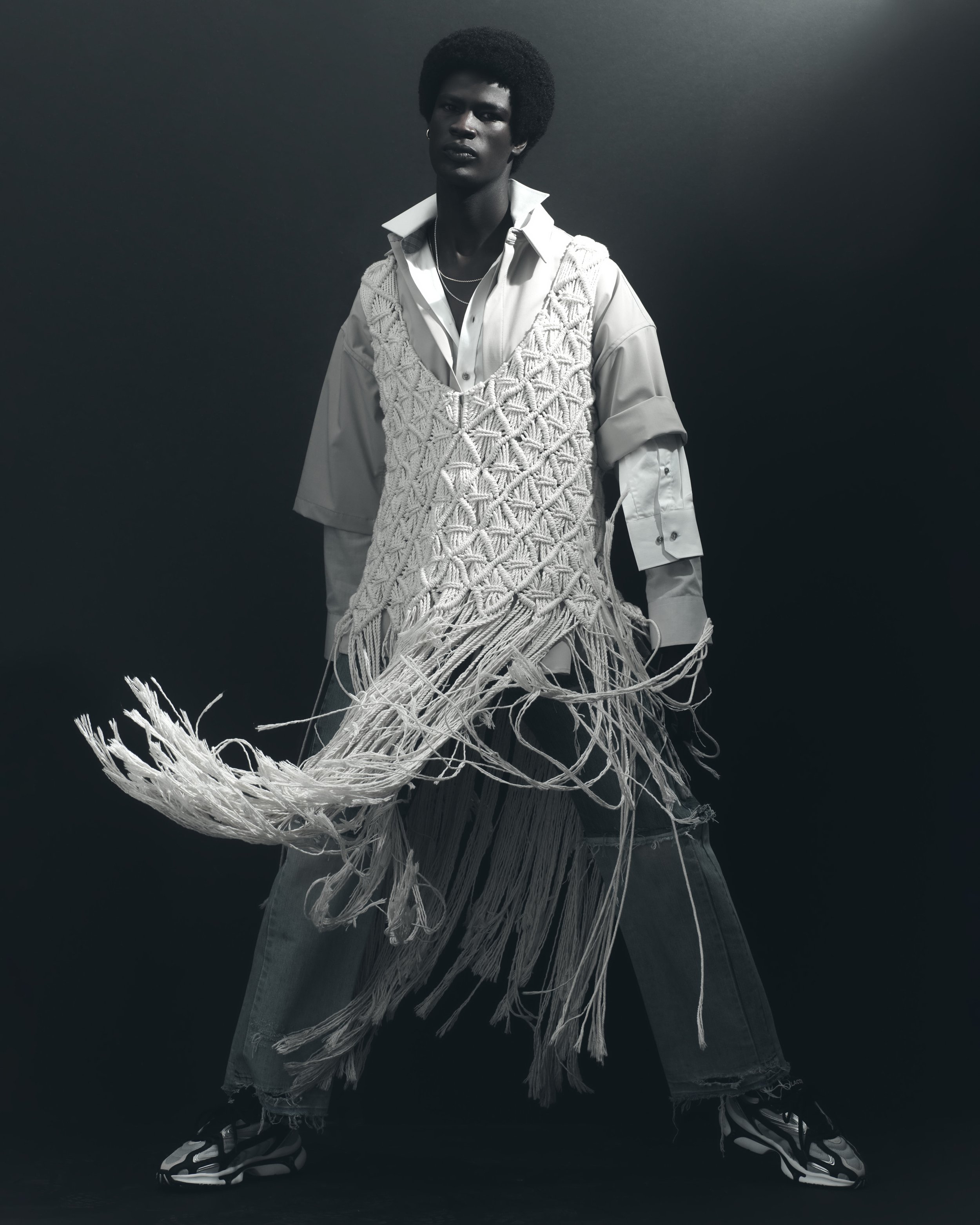 A person posing in a fashion photoshoot wearing a white shirt, layered with a large, knit, fringe vest, baggy jeans, and sneakers, against a dark background.