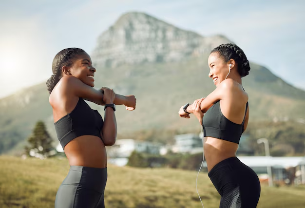 Two women in athletic wear smiling and stretching outdoors with a mountain in the background.