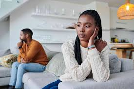 A woman sitting on a sofa, looking thoughtful, with a man sitting on the sofa behind her, in a modern living room.
