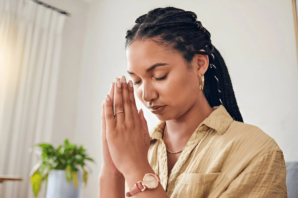 A woman with braided hair, closed eyes, and clasped hands in a prayer position, wearing a beige shirt and jewelry, in a peaceful indoor setting.