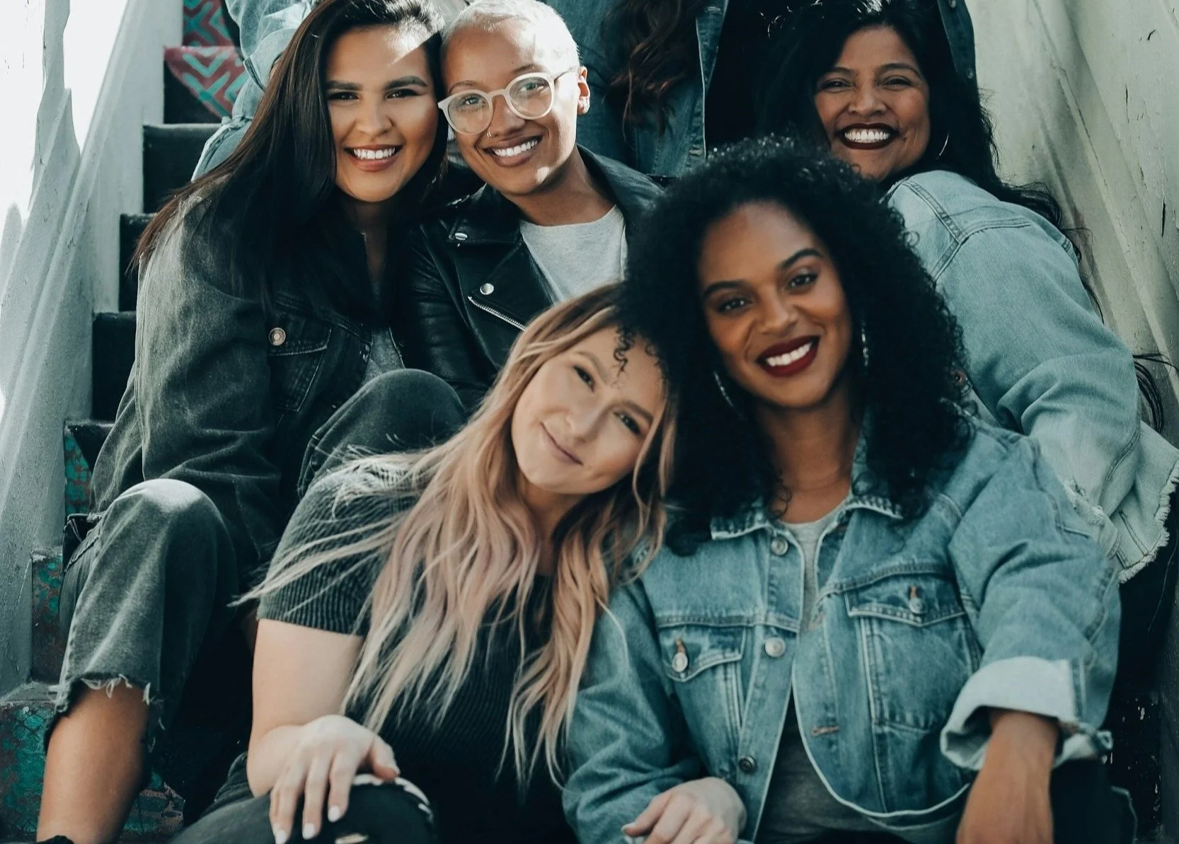 Group of five diverse women sitting on a staircase, smiling at the camera, wearing casual clothes and denim jackets.