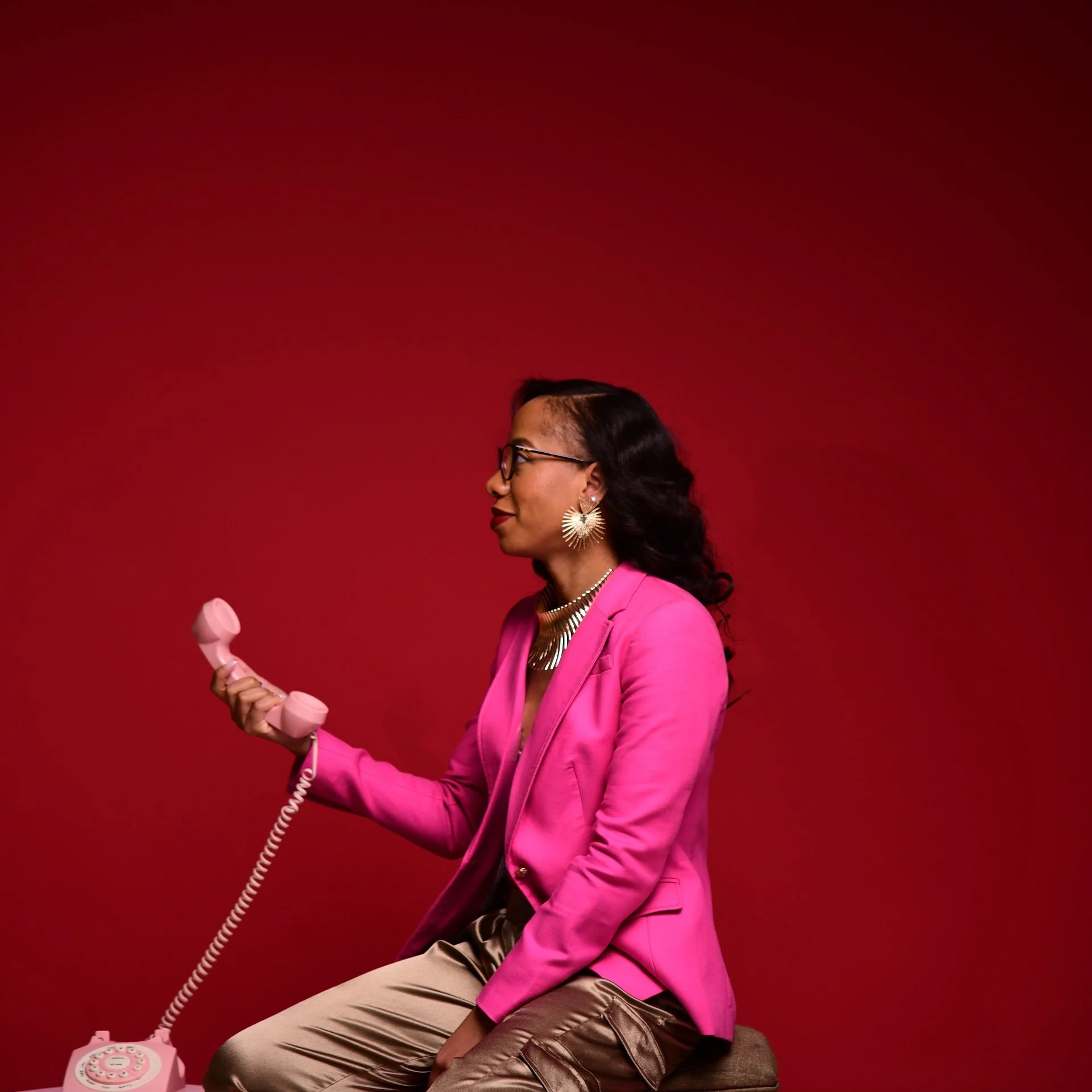 Woman in pink blazer and beige pants, wearing glasses and statement earrings, holding a pink rotary phone against a red background.