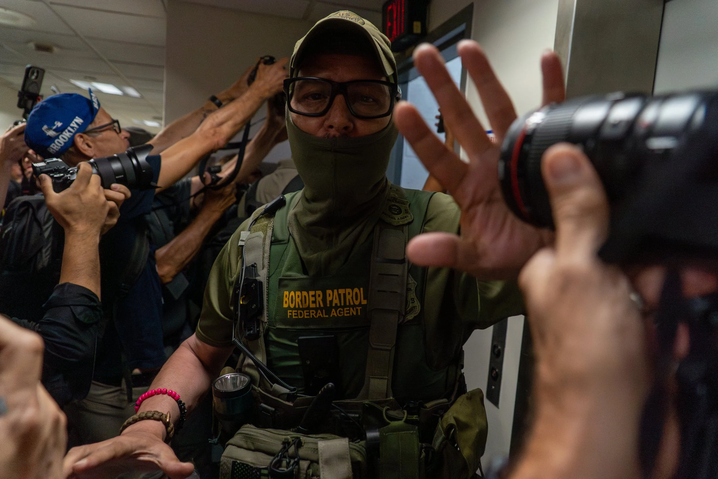 A Border Patrol agent lifts his hand toward a camera as press close in, documenting the tense walk to the stairwell. This moment—caught between flash and silence—captures the friction that follows nearly every detainment, as officers rush to remove t