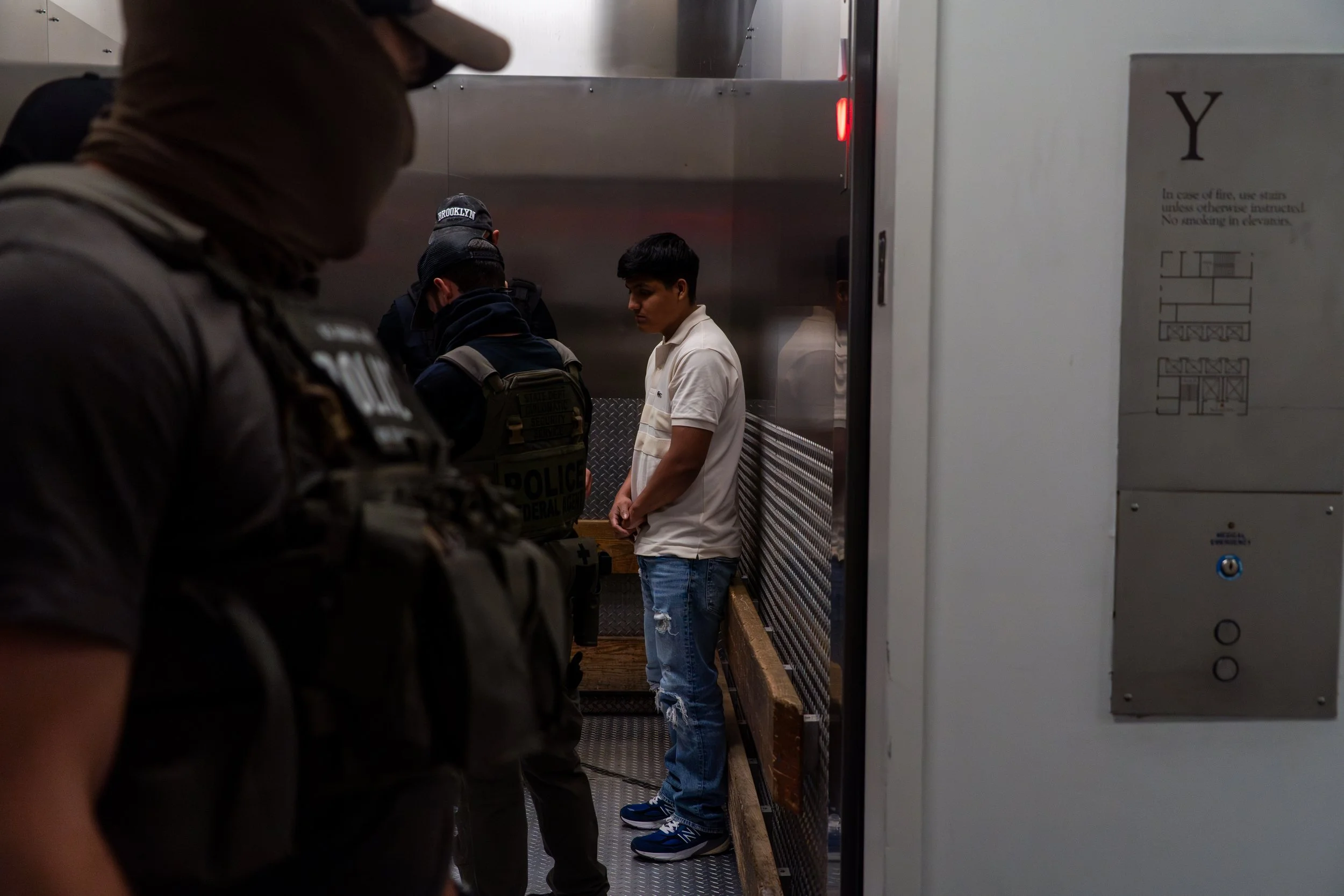 A young man stands alone in the back of a service elevator, surrounded by ICE agents in tactical gear. His hands are in front of him, his head slightly down. There is no struggle — just quiet compliance as the doors prepare to close.