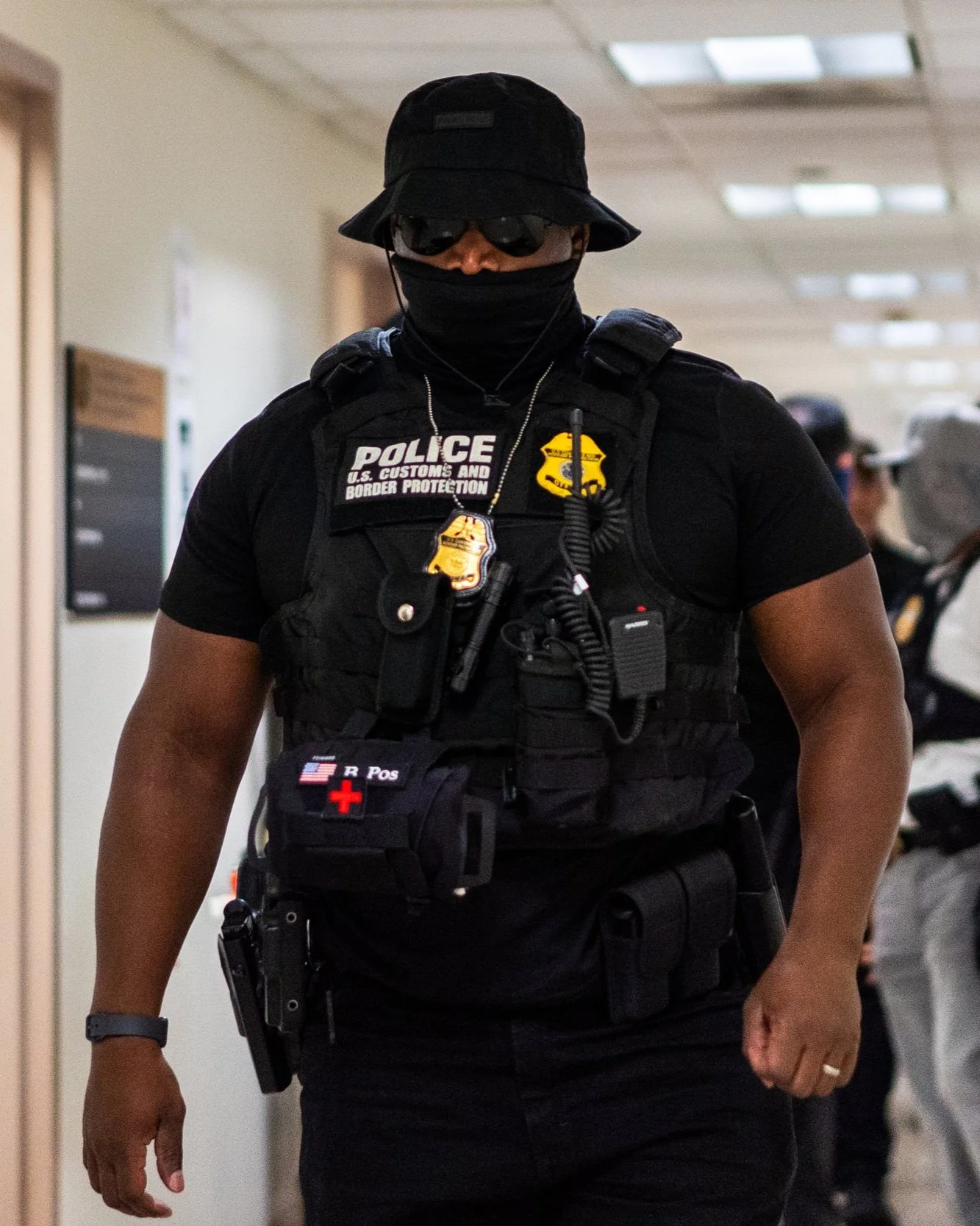 A U.S. Customs and Border Protection officer walks through the hallway in full tactical gear — face covered, vest armored, weapons and radio in place. His presence is unmistakable.