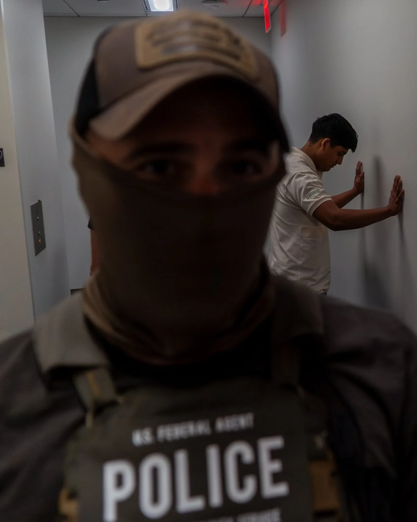 A masked ICE agent blocks the camera as a young man is ordered to face the wall. The tension in the hallway is palpable — arrests like this unfold quickly, quietly, without warning.