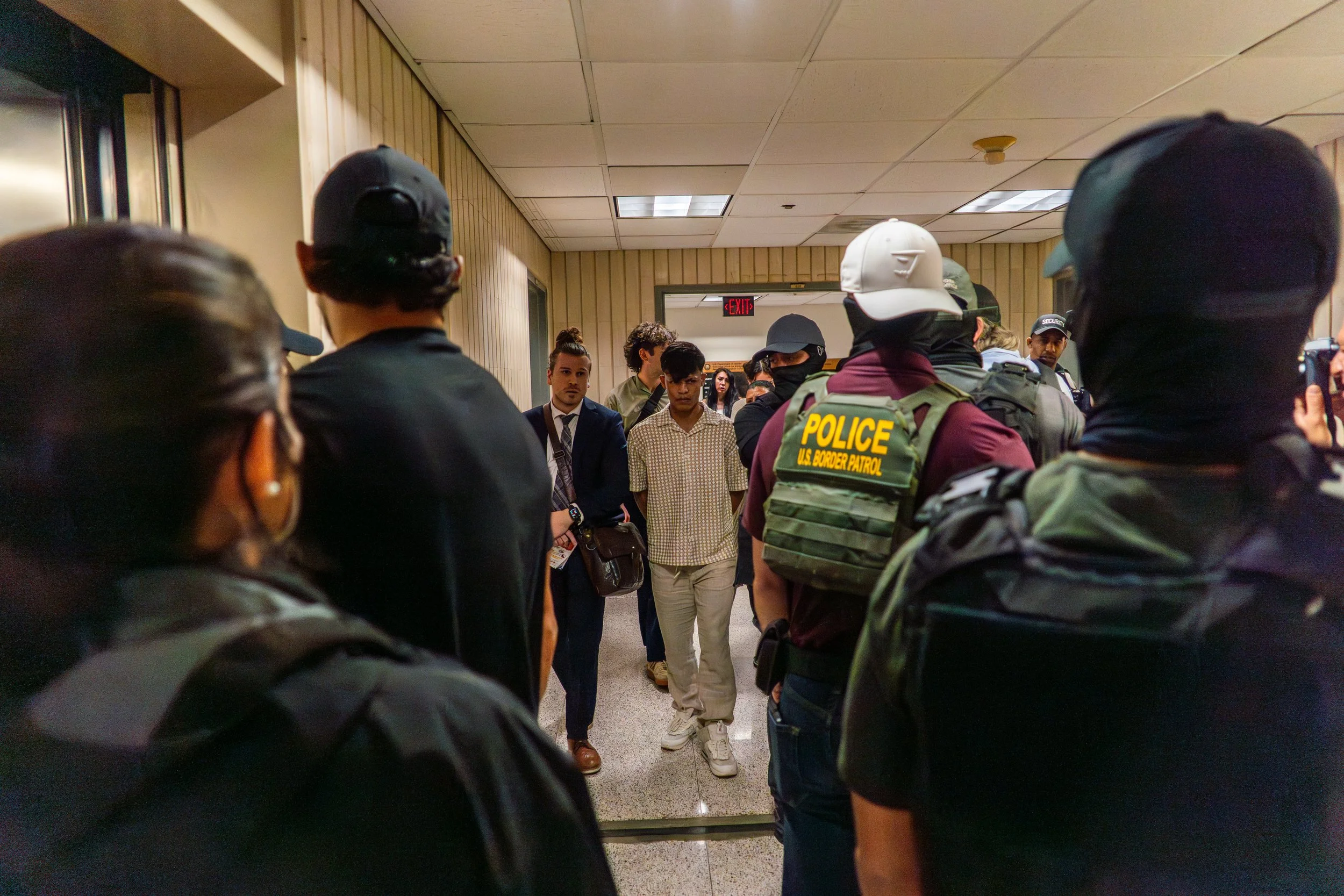 The boy at the center of this frame has just been detained by ICE. His mother, seen sobbing earlier while holding a baby, had no warning. He is now being led to the elevator— the beginning of a separation they weren’t prepared for.