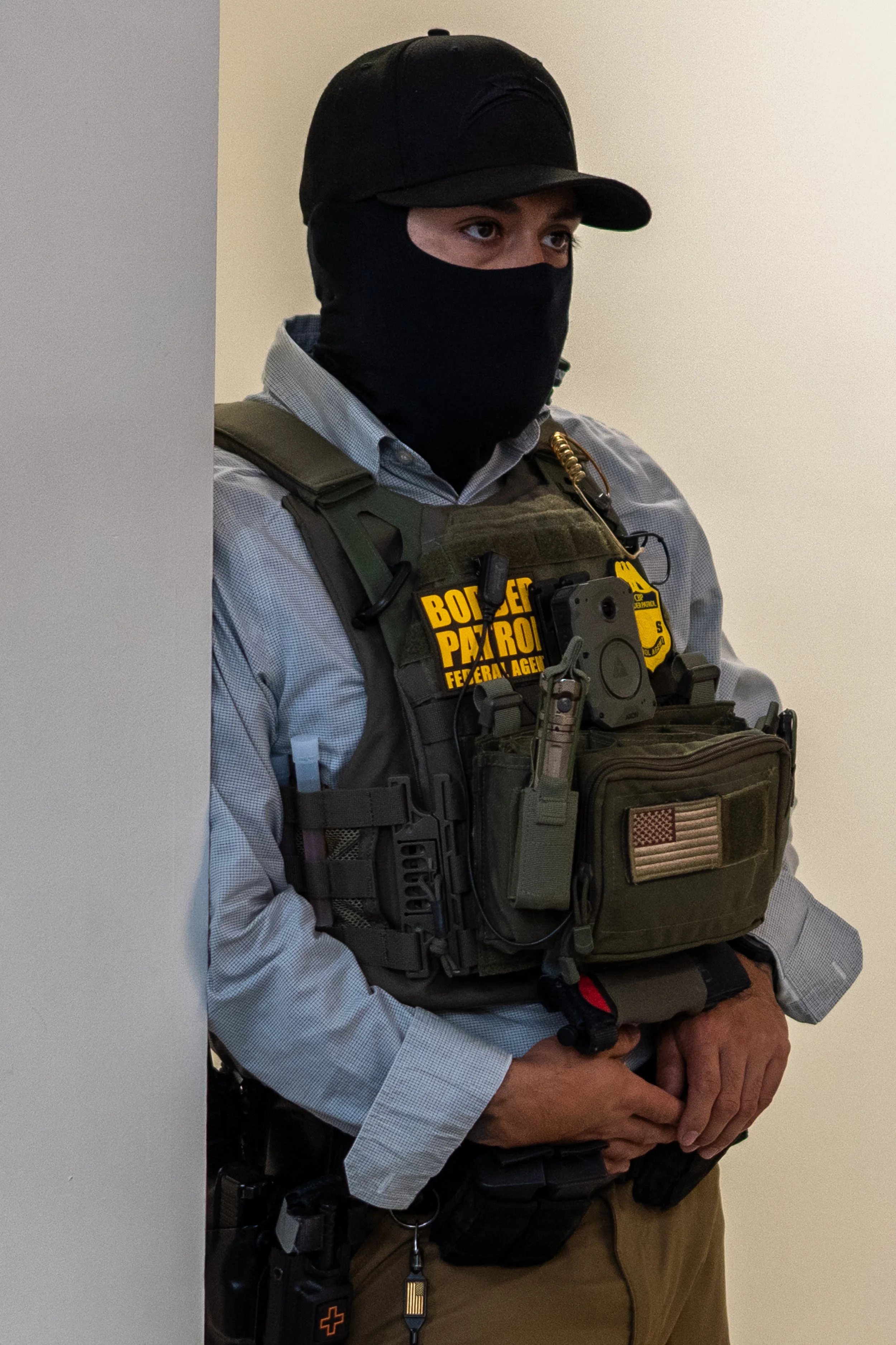 He stood mostly still, tucked against the wall just outside the courtroom. No name tag, no visible badge—only the black mask and tactical vest marked him as law enforcement.