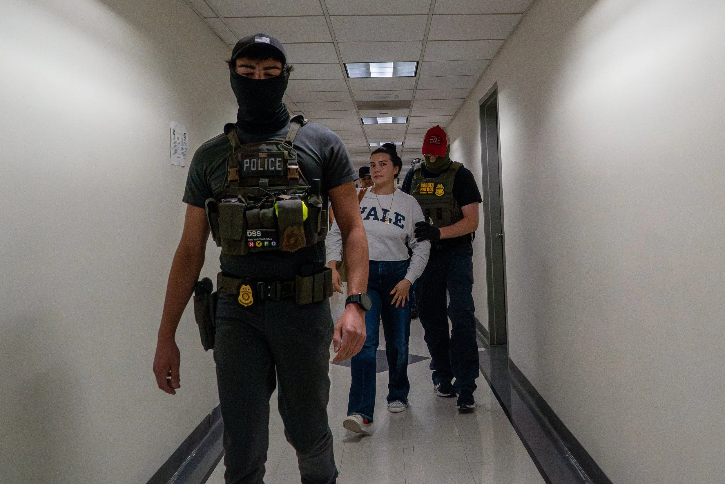 A young woman wearing a YALE sweatshirt is escorted down the hallway by ICE agents after her name is called. Her eyes are fixed forward, her body tense.
