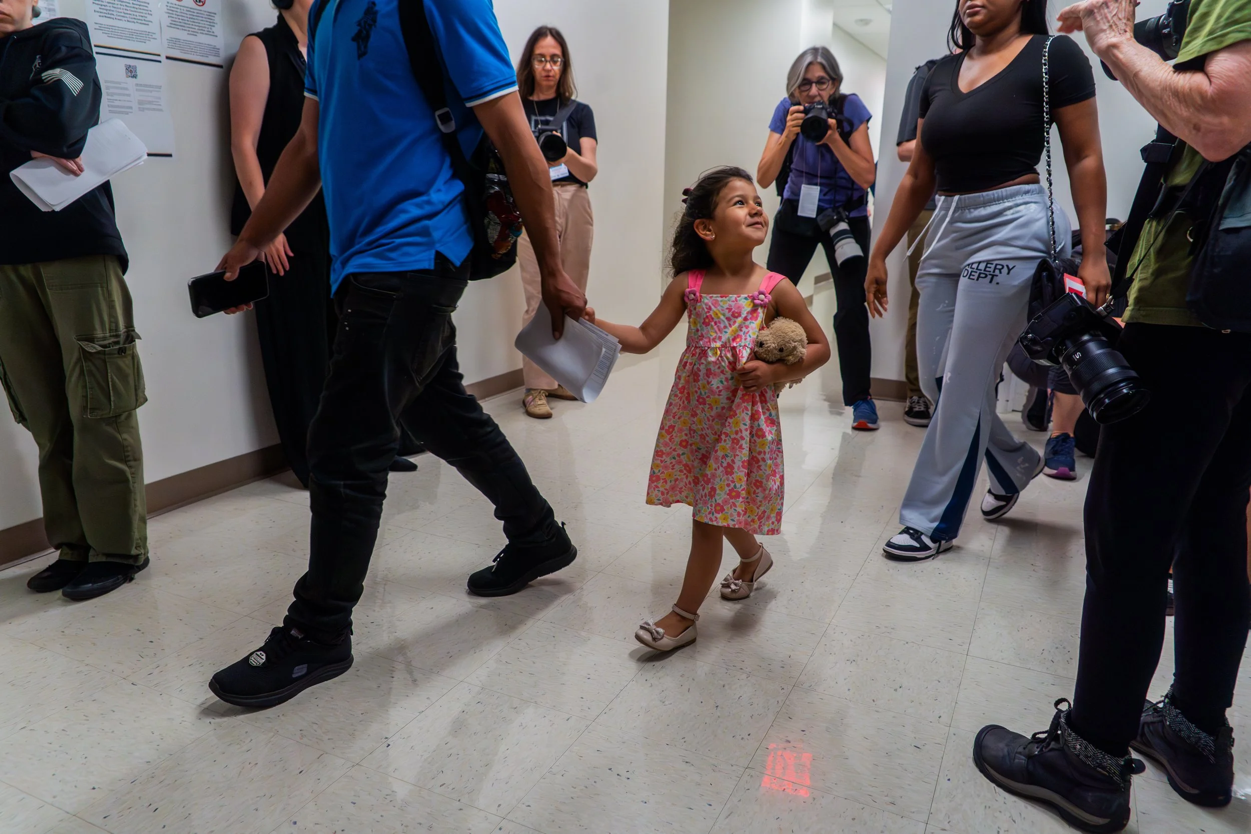 A little girl walks into immigration court holding a stuffed animal, smiling brightly as cameras follow.