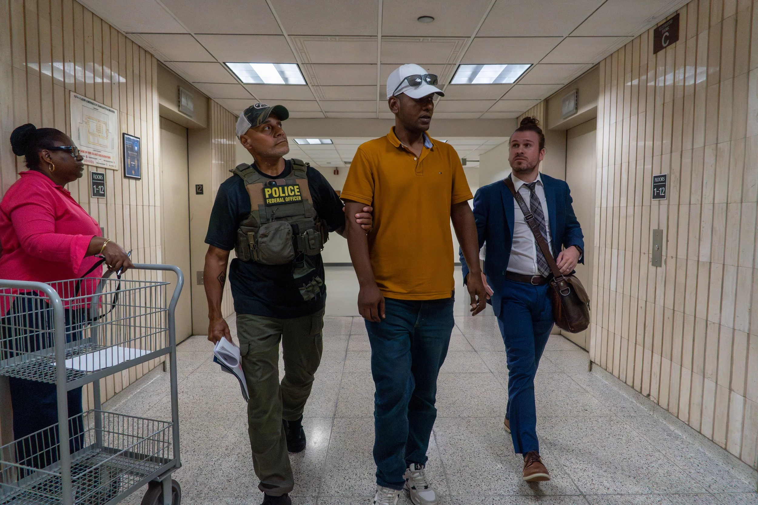 An ICE agent walks a man down the hallway after detaining him, his arm lightly gripped. A court clerk watches and an immigration attorney walks just behind. The system surrounds him — bureaucracy, legal counsel, enforcement — but none of it stops the
