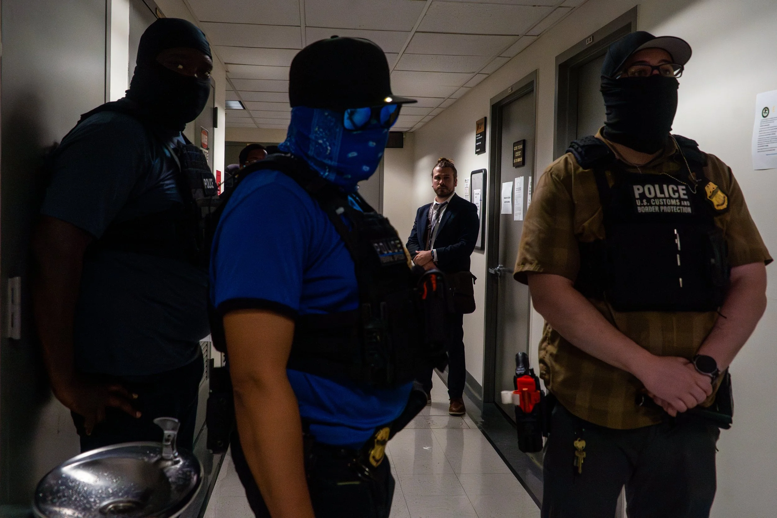 In the background stands immigration attorney Benjamin Remy, who returns week after week to advocate for those at risk of detention. In the foreground, ICE agents stand in silence.