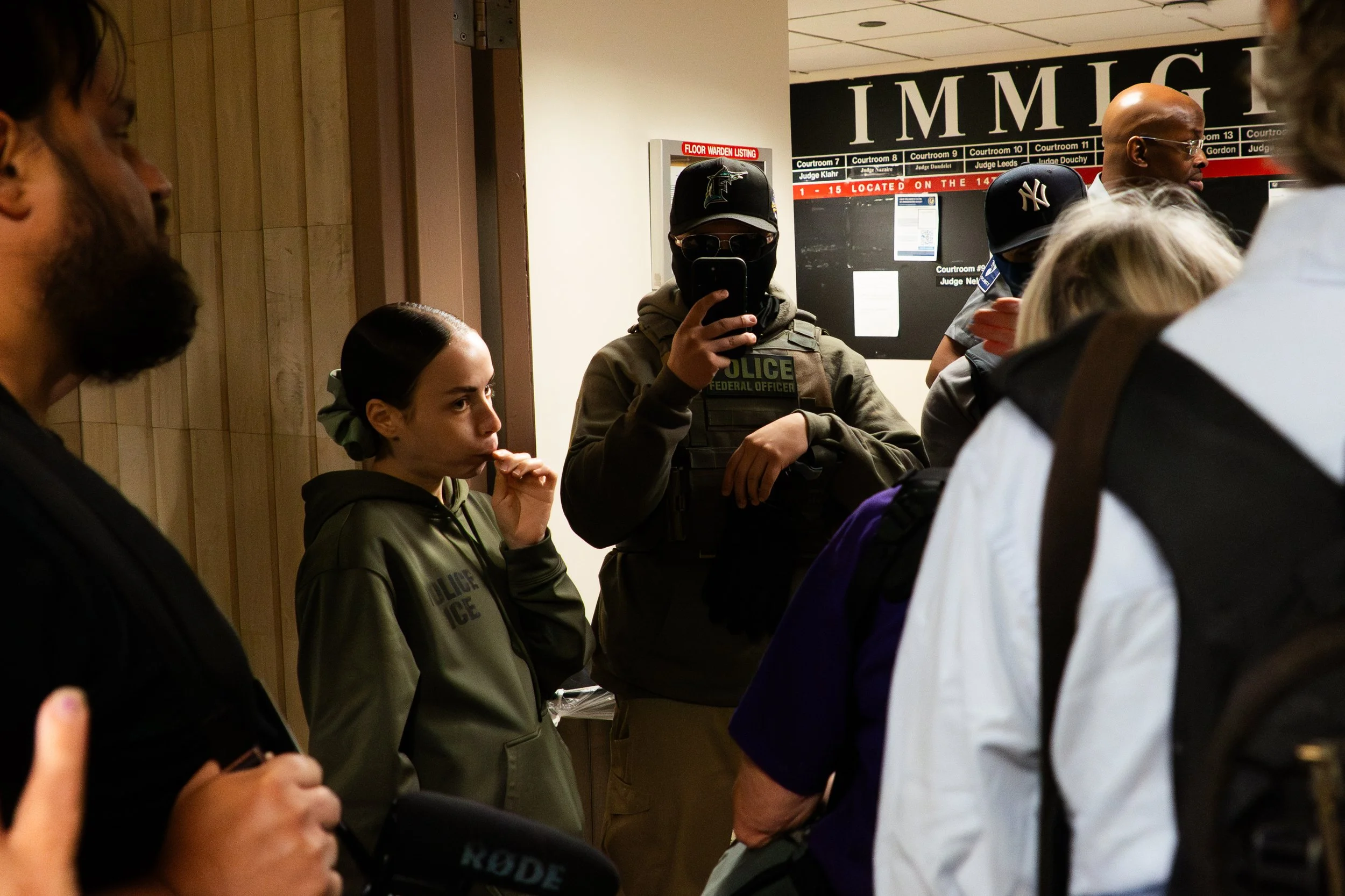 The head ICE agent — small in stature, dressed in a hoodie — sucks on a lollipop as she stands at the center of the hallway. Press, court observers, and federal agents move around her. The image is quietly jarring: casual, almost childlike, amid the