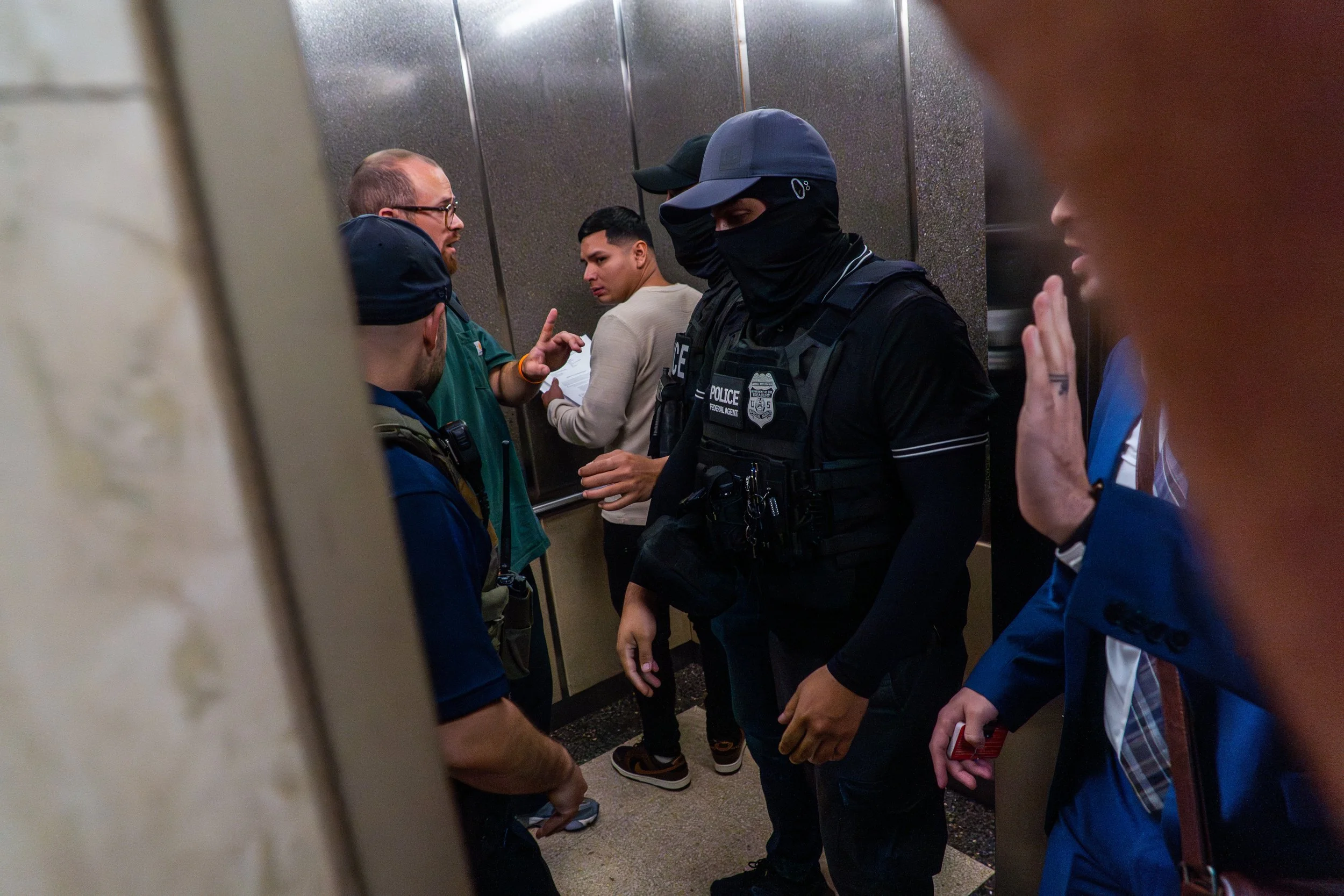 ICE agents step into the elevator with a man on their list and a court observer beside him. The observer raises his hand, mid-argument, trying to push back or bear witness.