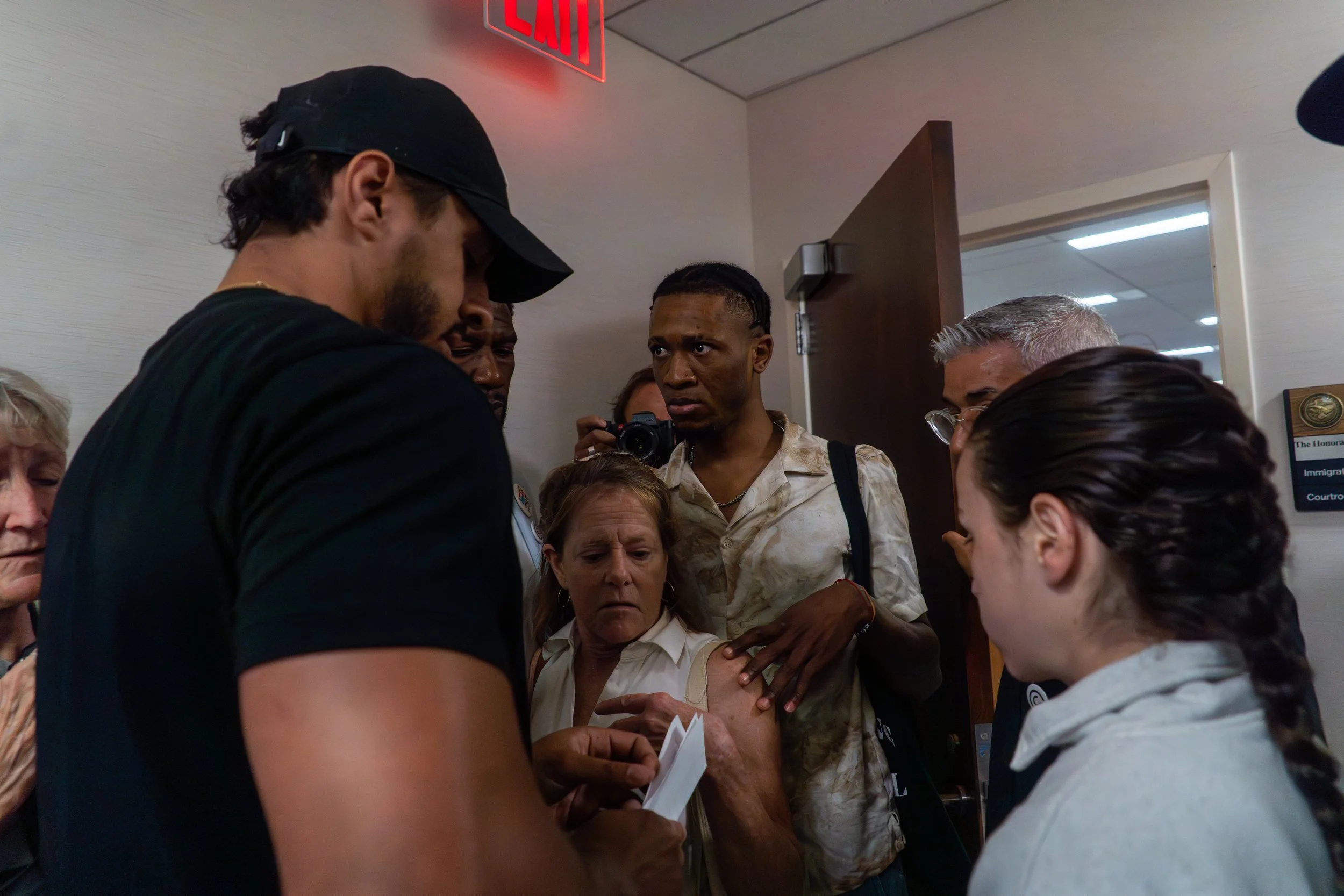 A man learns he’s about to be detained by ICE. His body tenses as the words sink in. In front of him stands a court observer, trying to shield him — speaking, questioning, protecting. But around them, the hallway tightens.