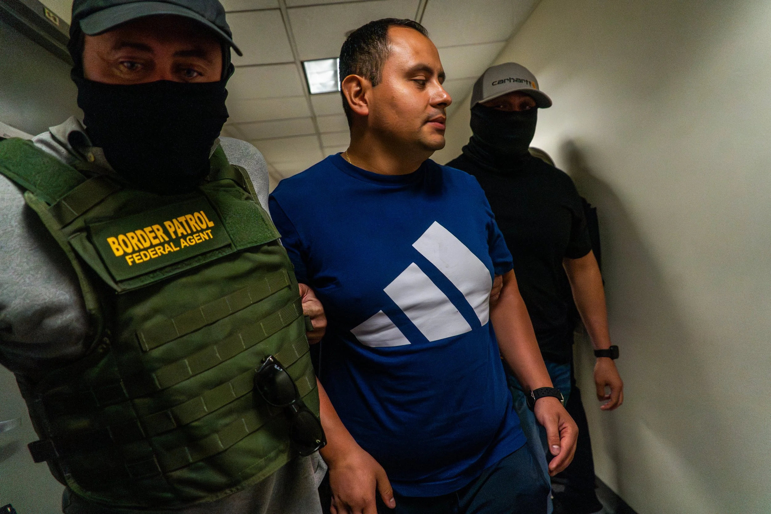 Two federal agents walk a man down the hallway after his immigration hearing. Escorted by officers in tactical vests and masks, he disappears around the corner—another name crossed off the list.