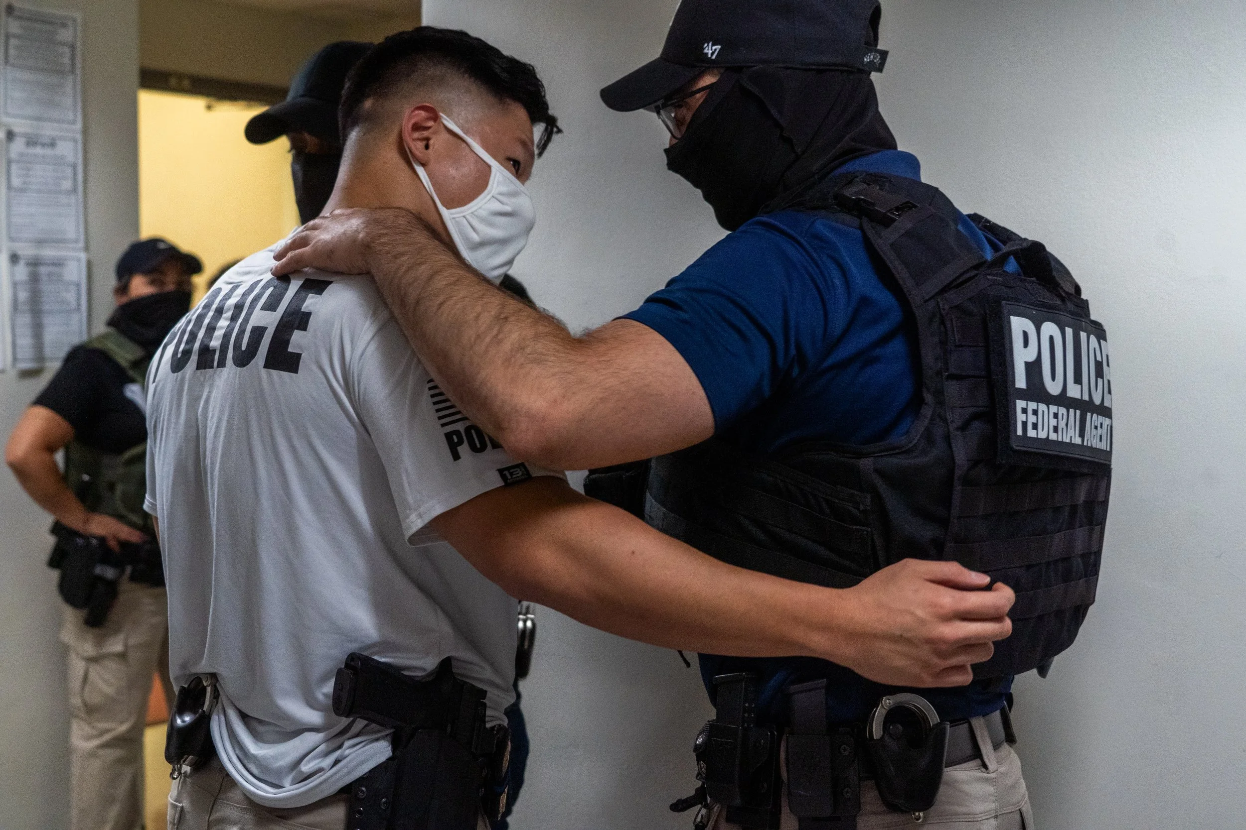 Two ICE agents share a brief moment in the hallway — a hand on the shoulder, words exchanged quietly.