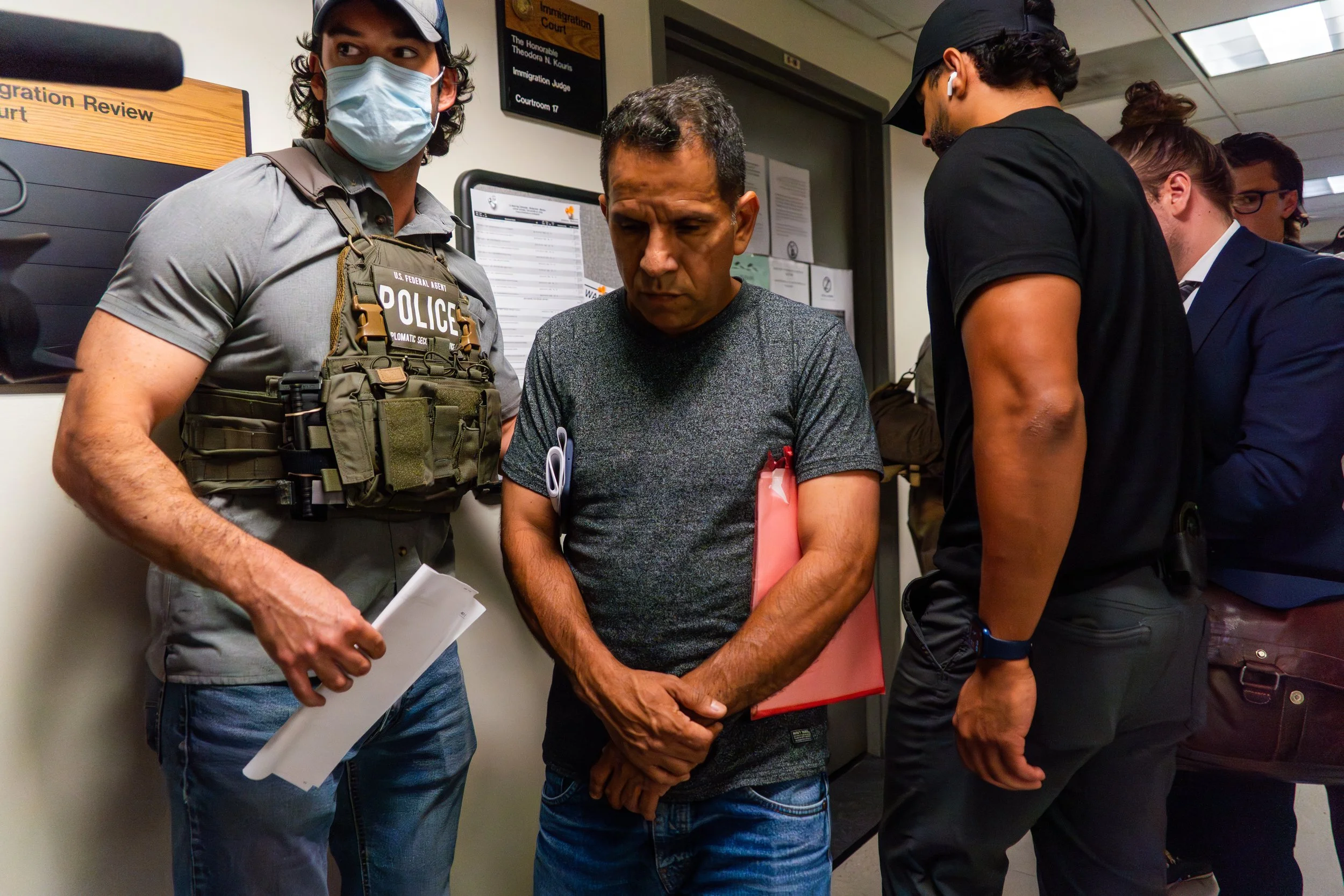 He has just been told there is a warrant for his arrest. Standing outside the courtroom, arms folded, eyes lowered, he does not protest — he simply goes still. The weight of that moment settles into his body as ICE agents surround him.