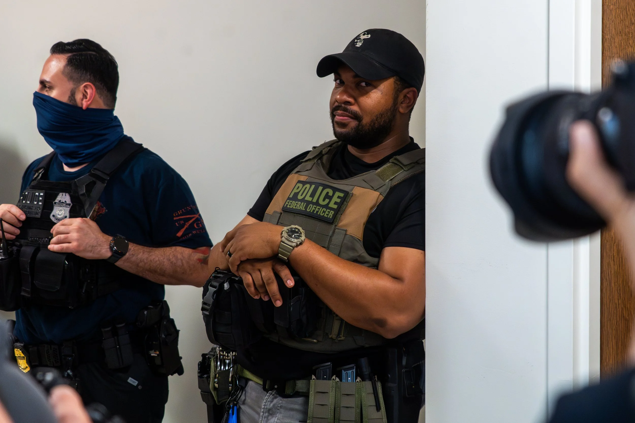 An ICE agent leans casually against the wall, smiling as a press camera captures the moment.