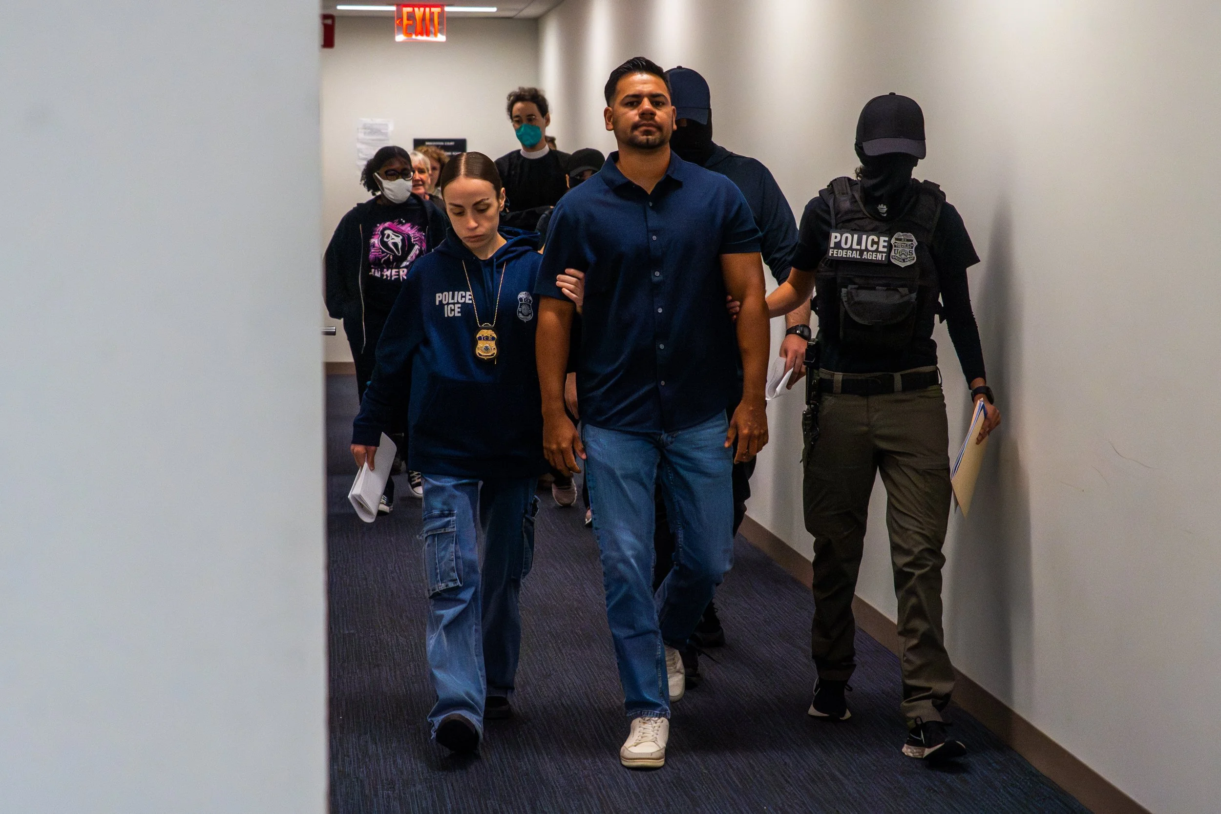 A man is flanked by three ICE agents as he is escorted down the hallway — one at each arm, another following behind. The agents wear hoodies and tactical gear, their expressions obscured, while his is clear: defiant, composed.