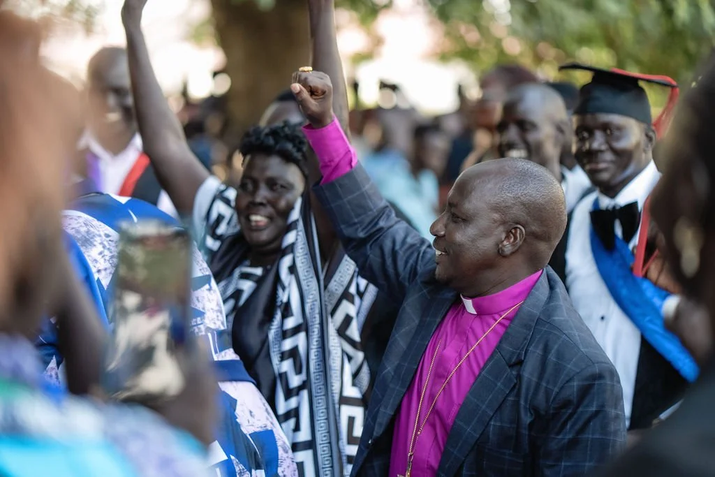Crowd of people outdoors with hands raised against a backdrop of trees and sunlight.