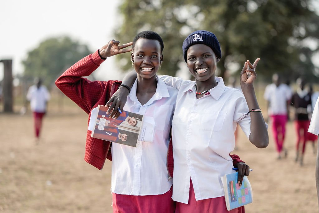 Person holding a pen with the text 'Hope Secondary School, Lietnhom' on it, wrapped in plastic, with a blurred figure in a white shirt and red tie in the background.