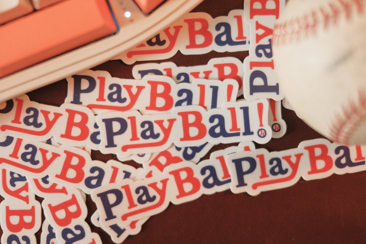 Pile of stickers sitting next to a baseball. The stickers read Play Ball! in a fun hand-lettered font.
