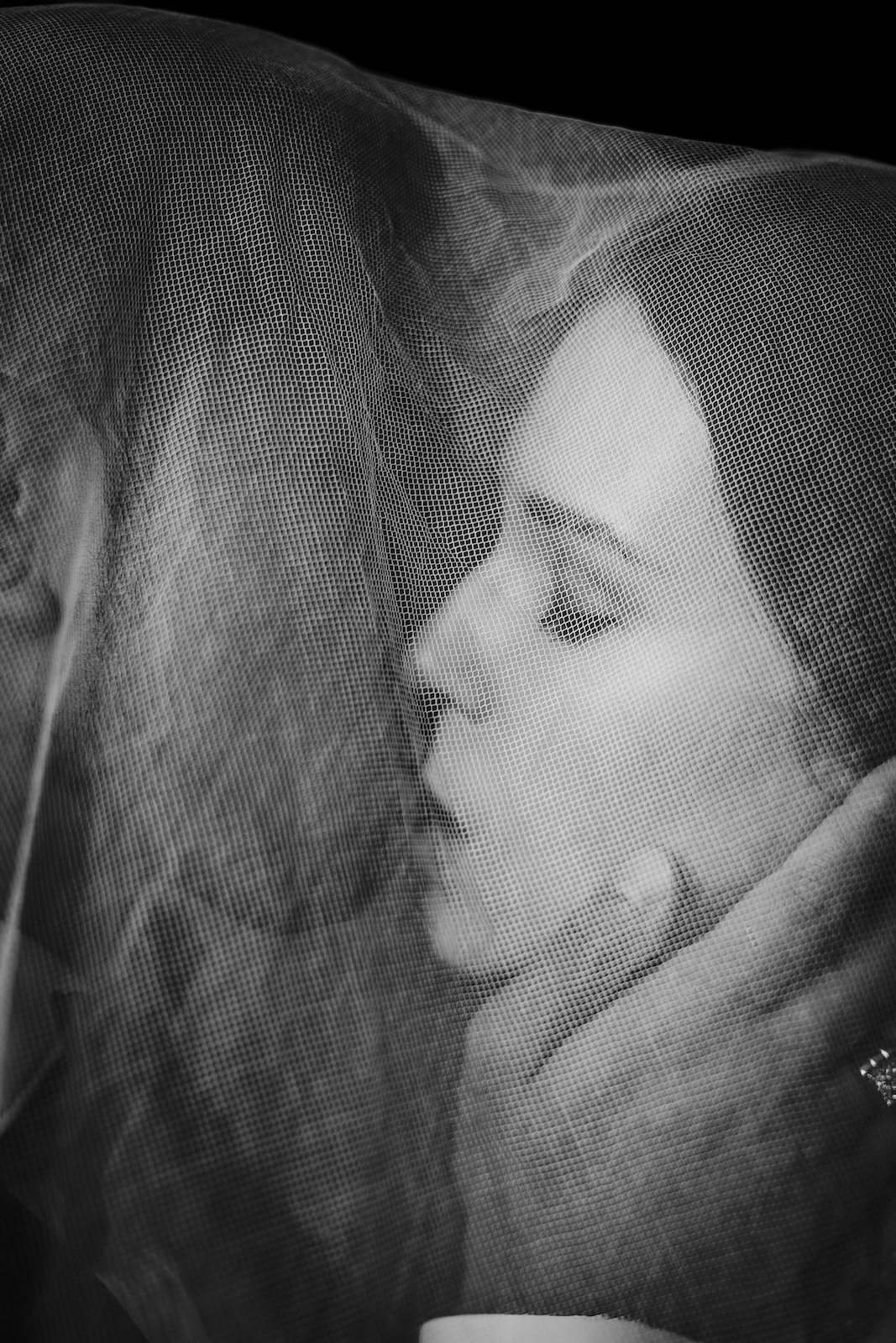A monochrome image of a woman behind a mesh or netting, with her hand near her face, creating a silhouette effect.