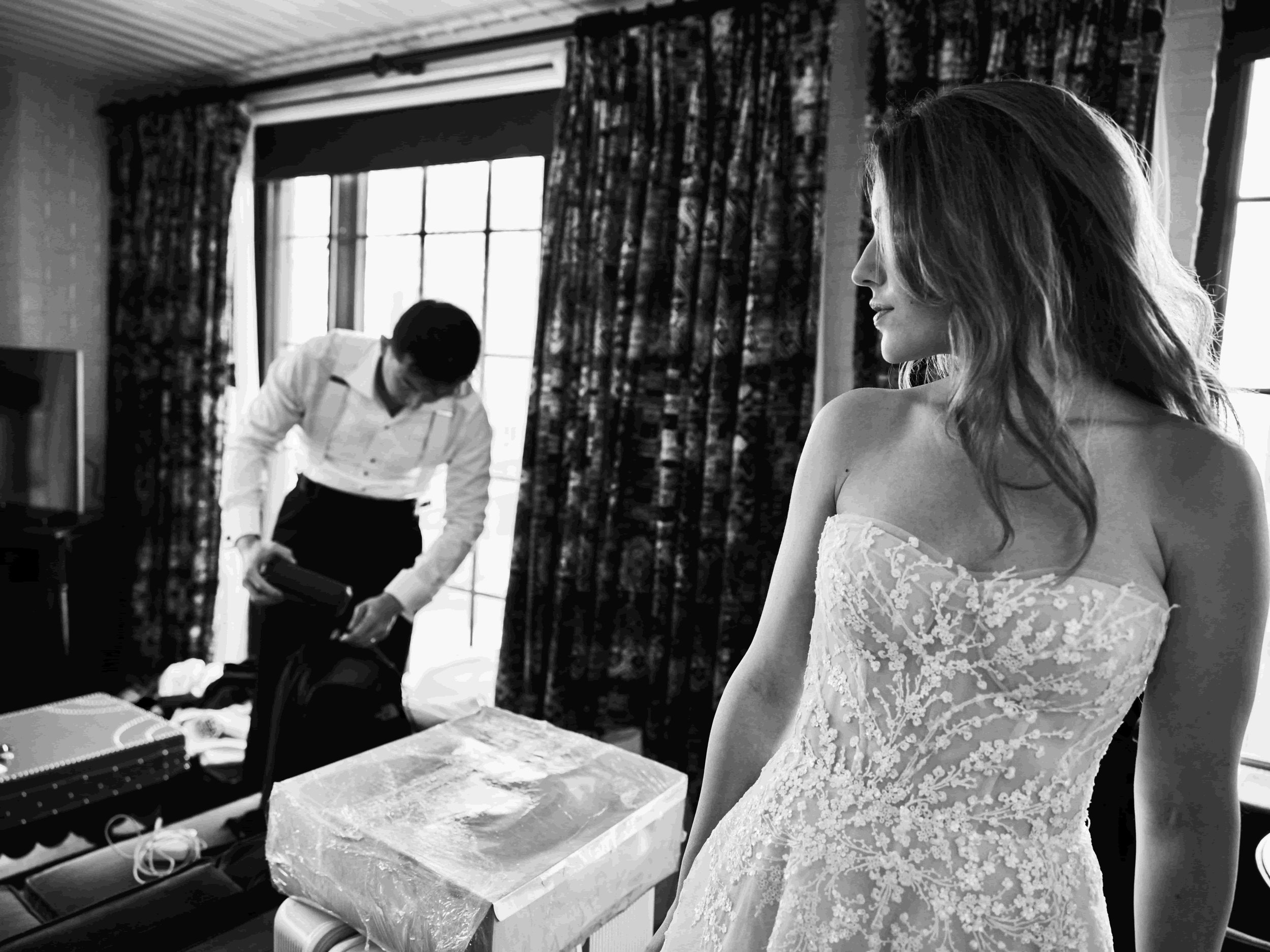 A woman in a wedding dress looking to the side in a dimly lit room, with a man in the background packing a bag near a window.