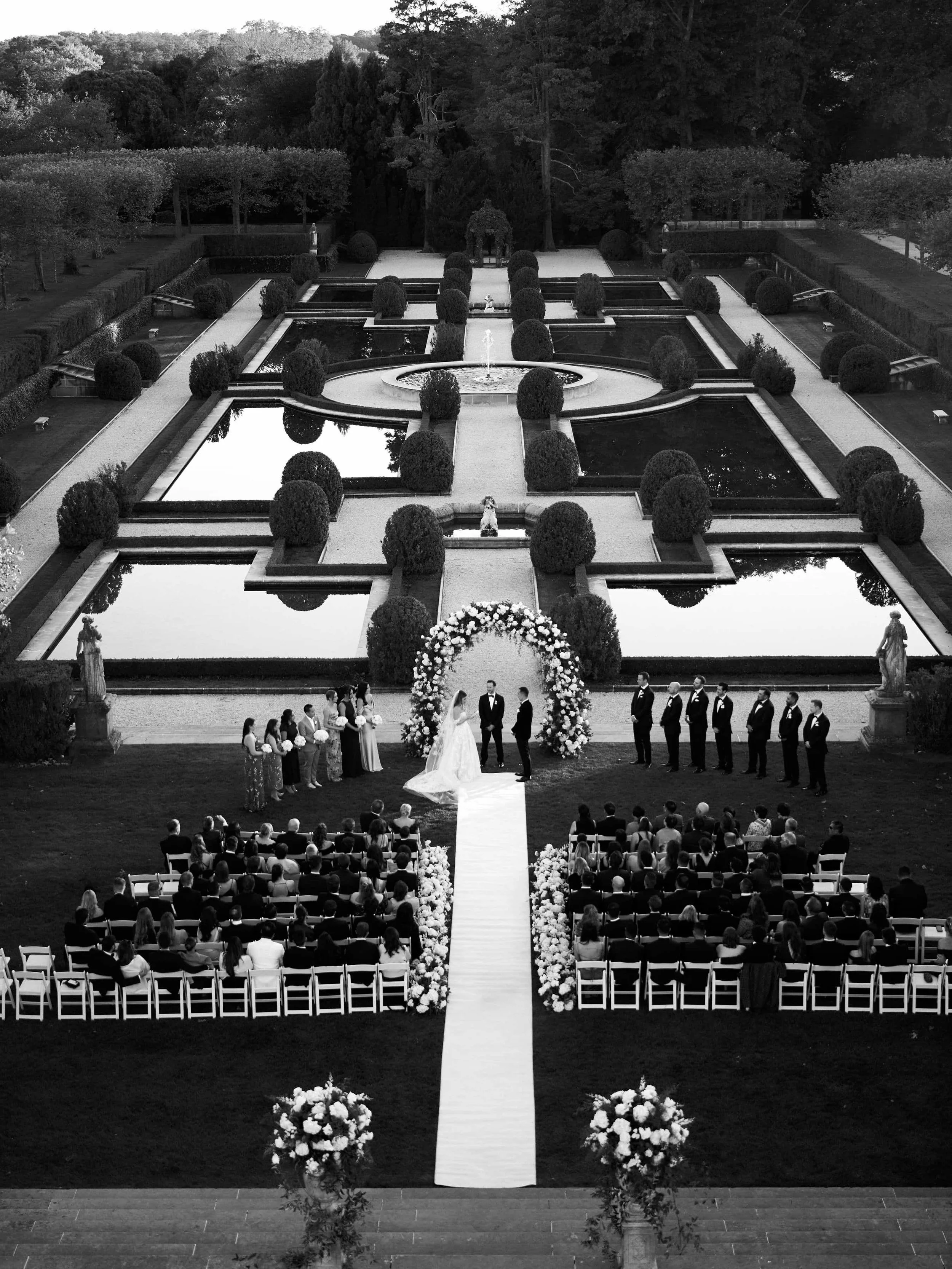 A black-and-white photo of an outdoor wedding ceremony taking place in a formal garden with water features, statues, and trees in the background. The bride and groom stand under a floral arch, surrounded by wedding party and guests.