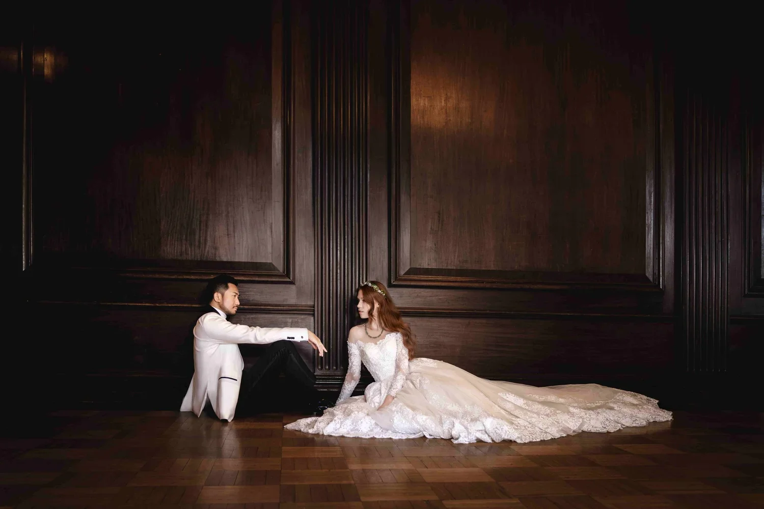 A bride in a white wedding dress and a groom in a white tuxedo sitting on a wooden floor in front of a dark wood-paneled wall, gazing at each other.