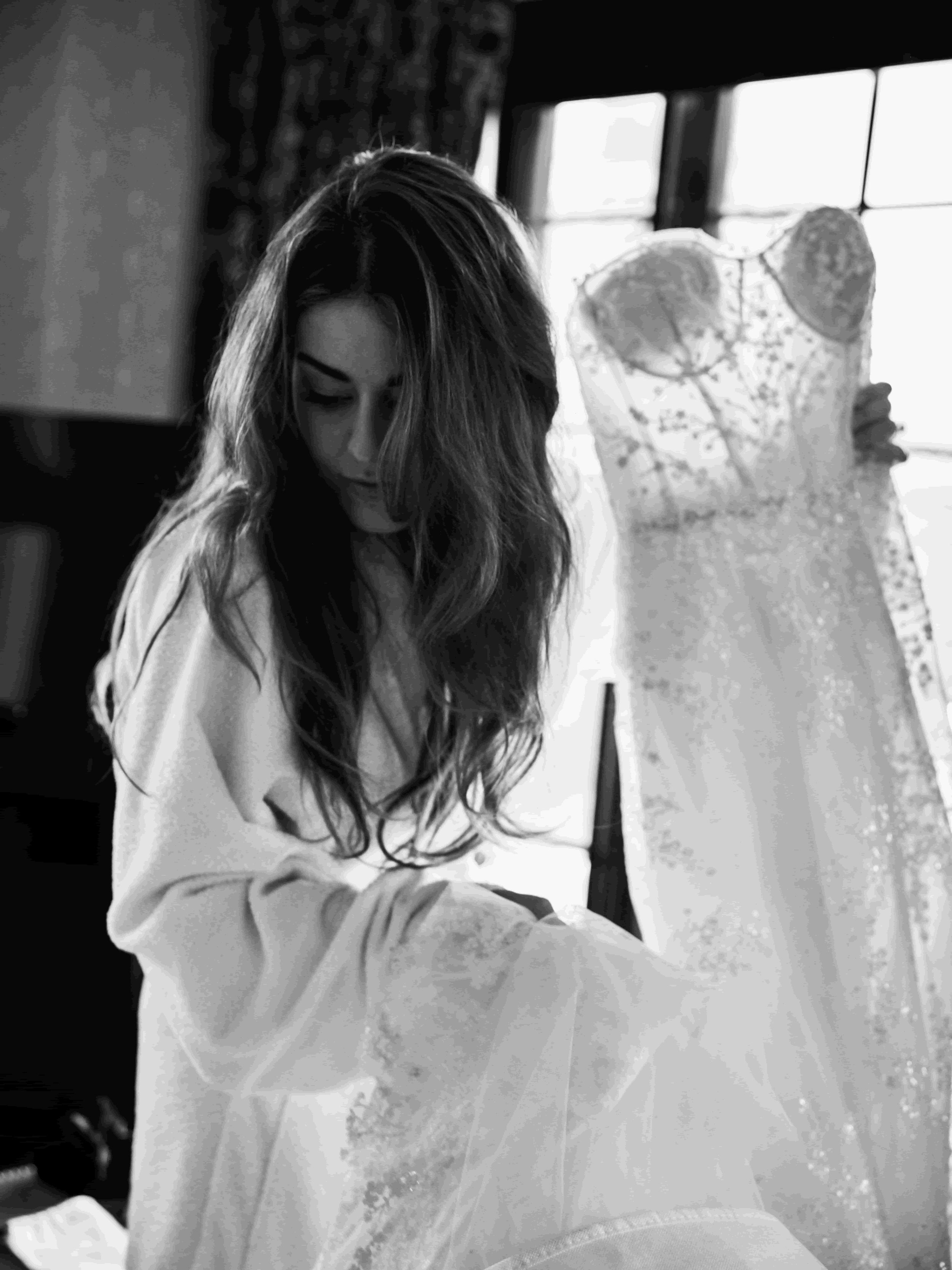 A woman with long wavy hair looking down in a room, with a wedding dress on a dress form in the background.