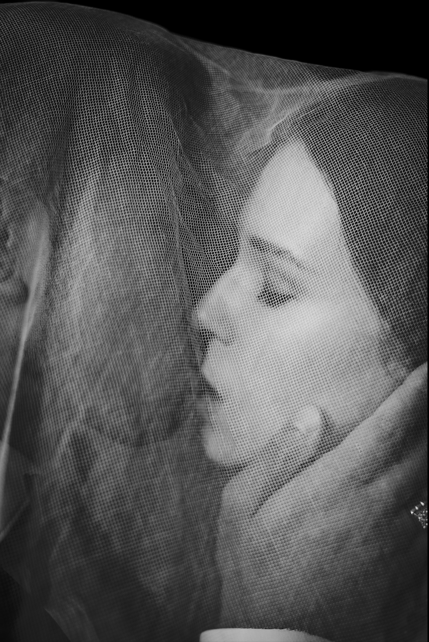 Close-up of a woman with closed eyes, behind a sheer veil, touching her face gently.