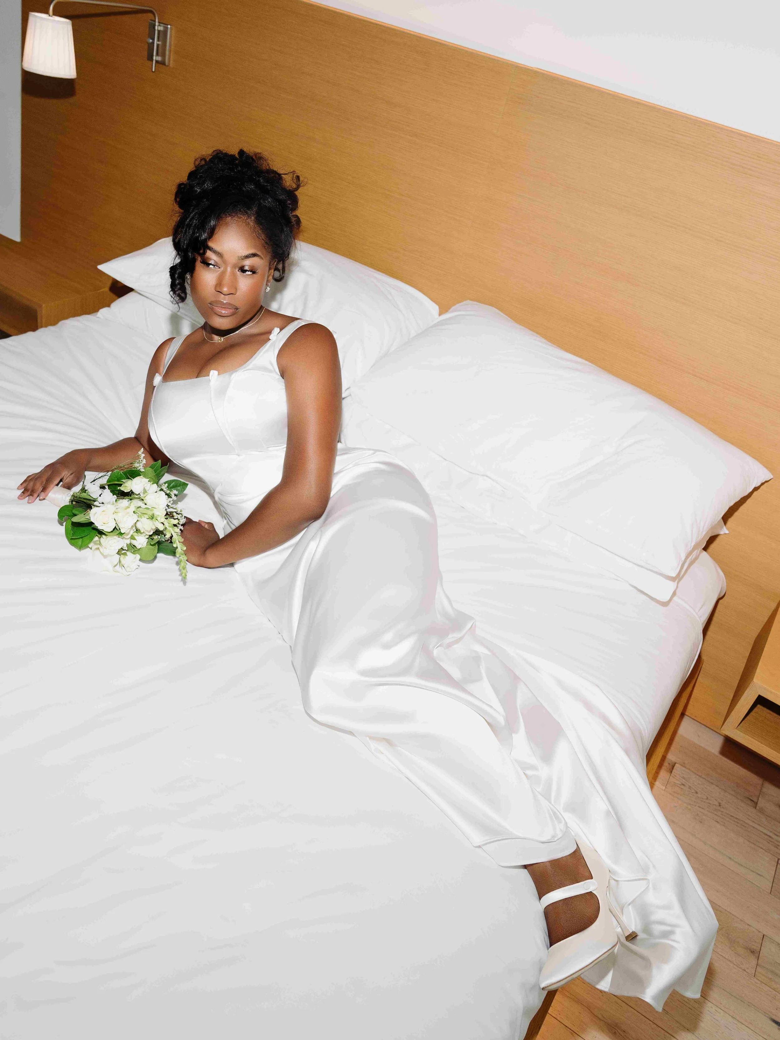 A woman in a white dress lying on a bed with white sheets and pillows, holding a small bouquet of white flowers.