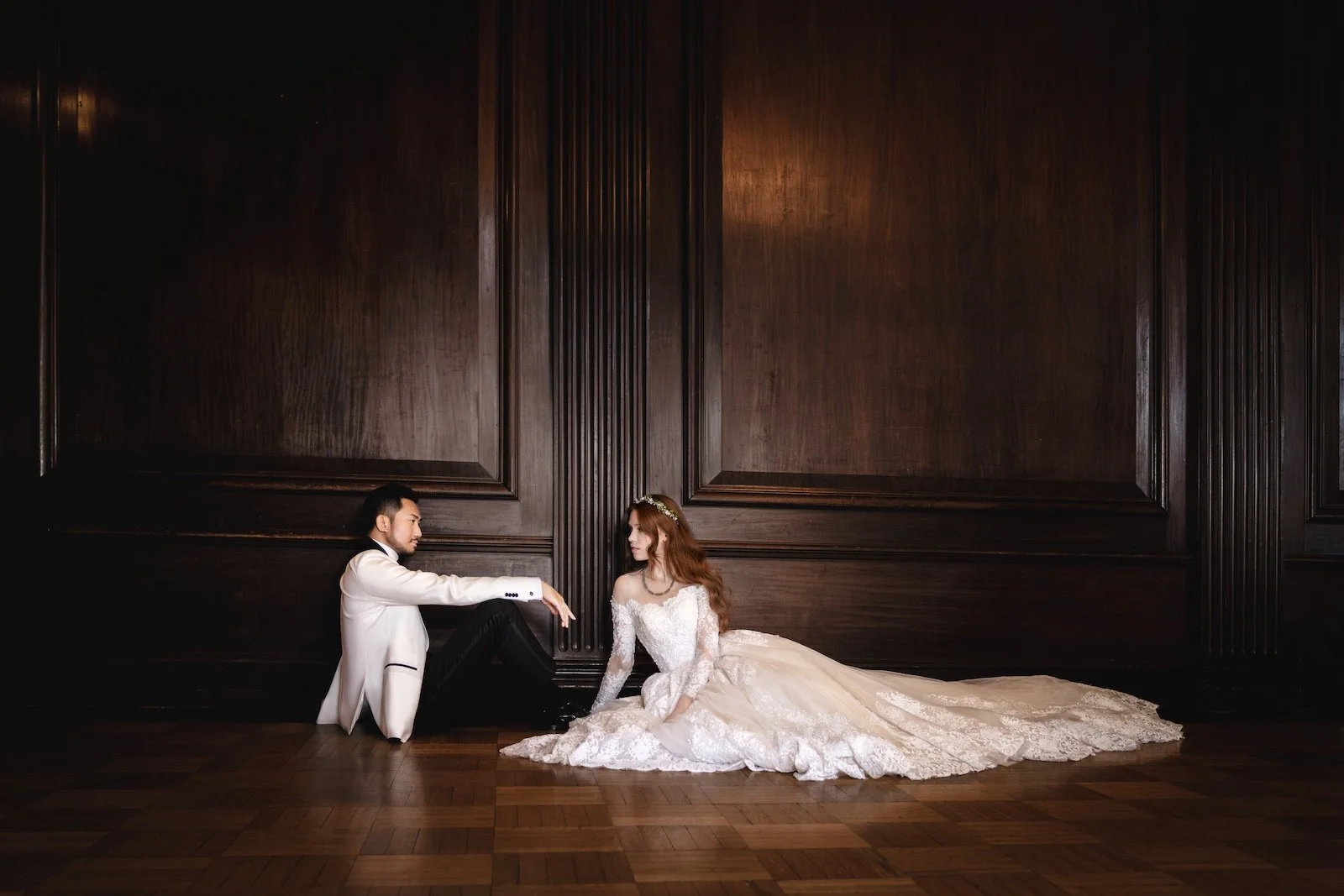 A bride and groom in wedding attire sitting on the wooden floor in front of a dark wood-paneled wall, facing each other with the groom extending his arm to hold the bride's hand.
