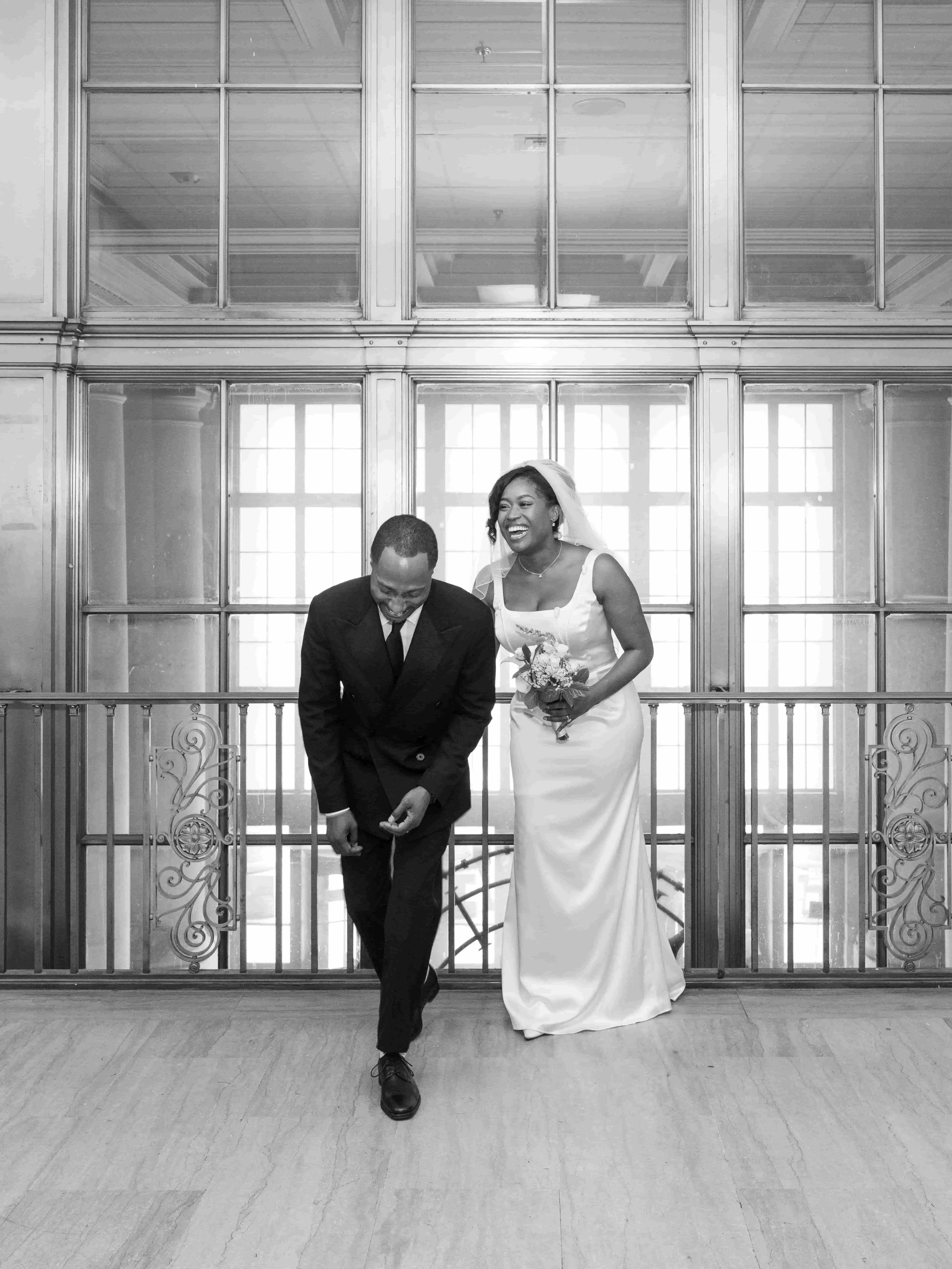 A bride and groom on their wedding day, laughing and walking together in an elegant, indoor setting with large window panels behind them.