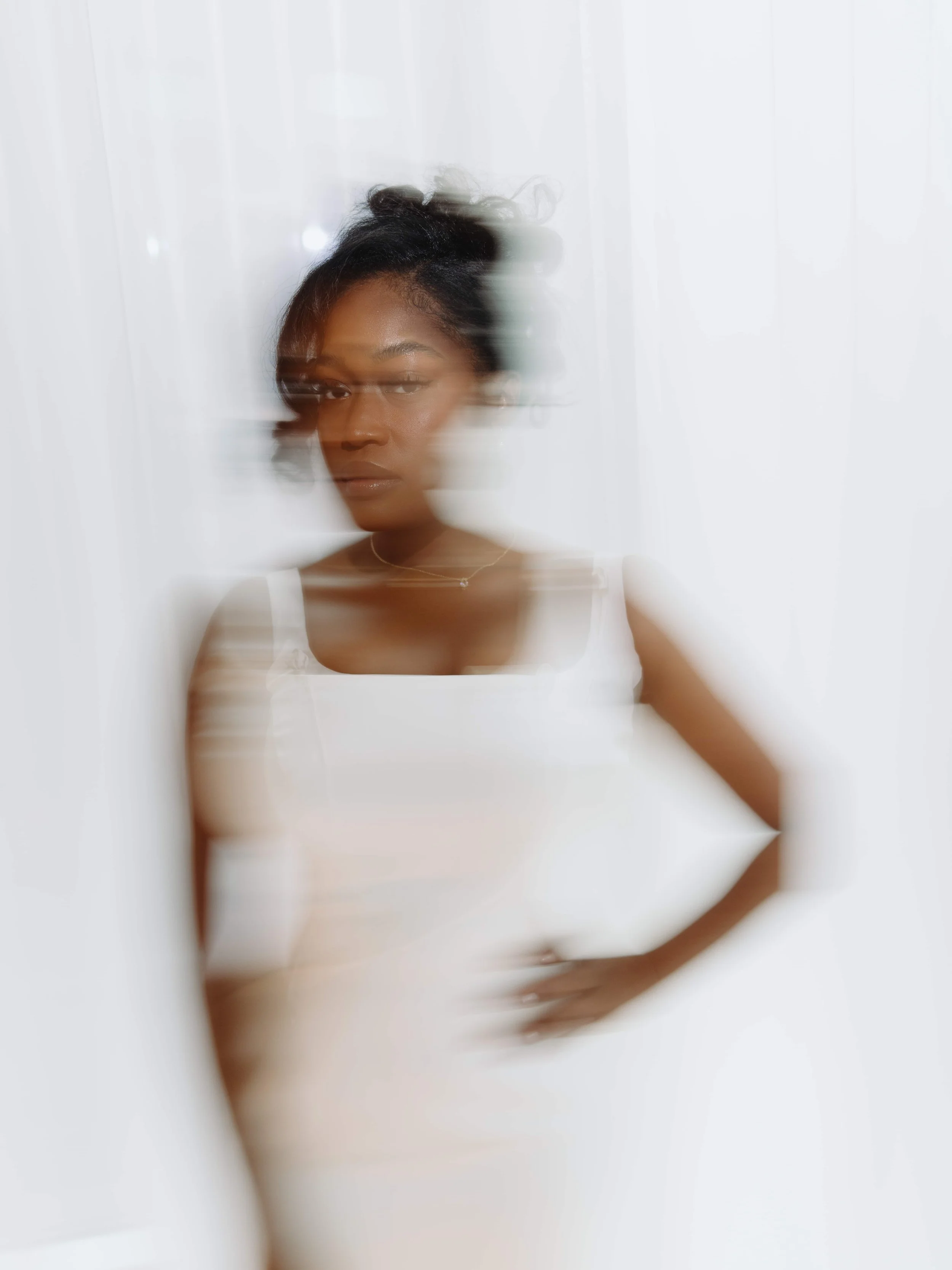 A woman with shoulder-length dark hair and a serious expression wears a white top and a delicate necklace, standing in front of a white background with motion blur effects.