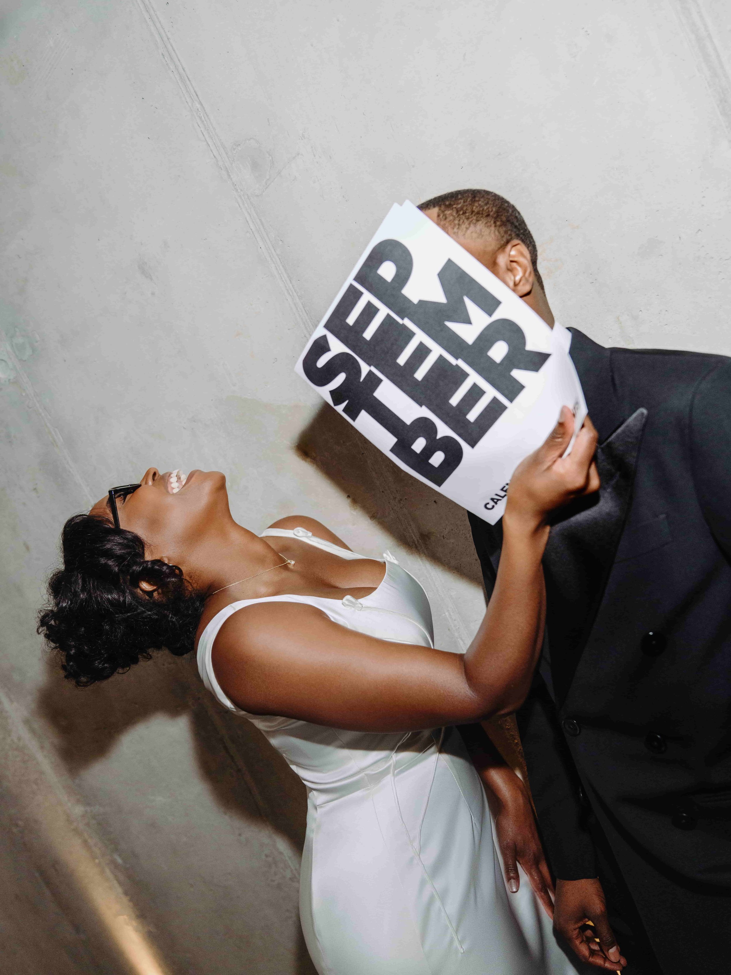 A bride in a white dress laughs while holding a protest sign near a man in a suit whose face is obscured by the sign.