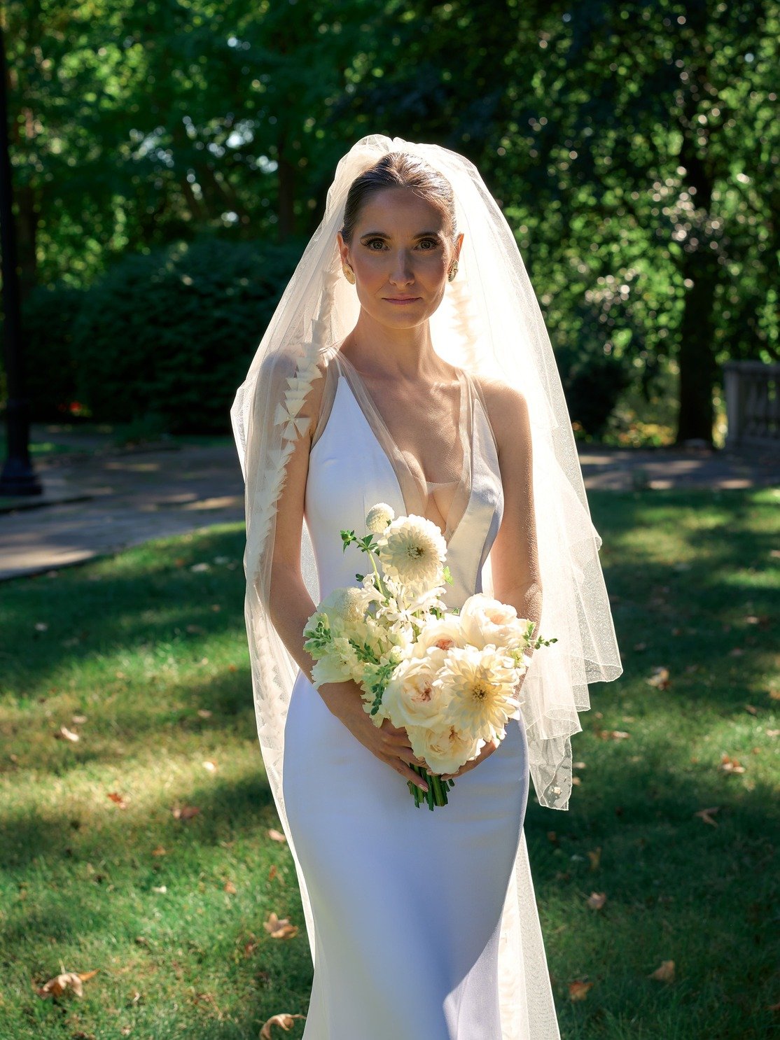 A bride stands outdoors on a sunny day, wearing a white sleeveless wedding gown and a sheer veil. She holds a bouquet of white flowers and looks directly at the camera with a calm expression, surrounded by green trees and grass.