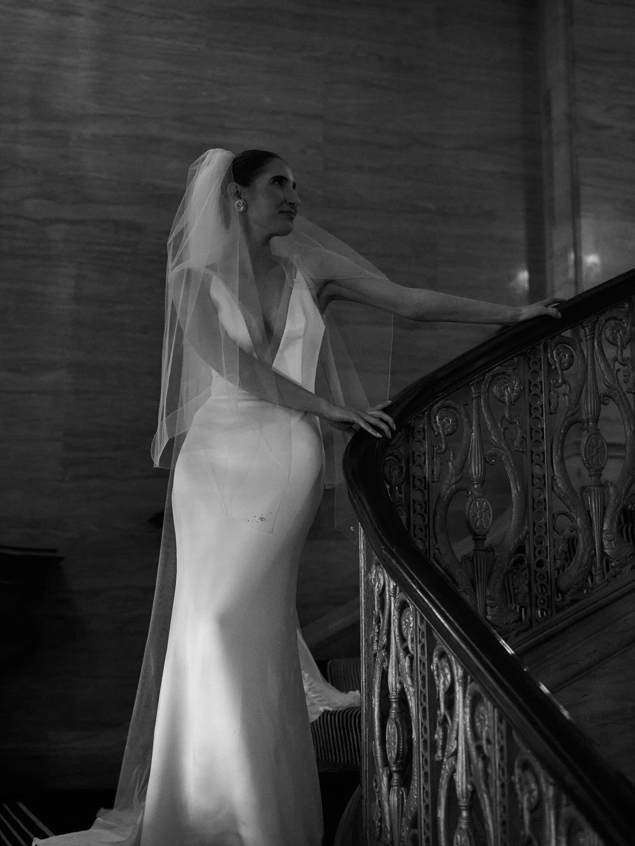 Black and white photo of a bride in a long gown and veil standing on a staircase.